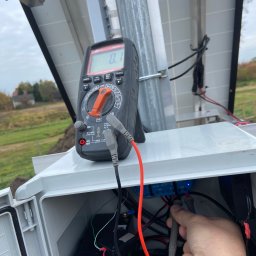 POLTHOM Paweł Kietla - Pomiar napięcia instalacji fotowoltaicznej multimetrem. Widoczne panele, skrzynka z okablowaniem i akumulator. Praca w toku, pomiar parametrów.