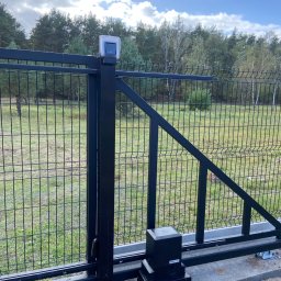 POLTHOM Paweł Kietla - Czarna, metalowa brama przesuwna z automatyką, widoczna na tle zieleni i drzew. Brama z siatki, z napędem i lampą ostrzegawczą. Ujęcie z dołu, podkreślające mechanizm.