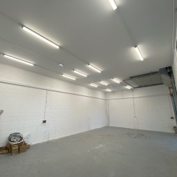 POLTHOM Paweł Kietla - Wnętrze hali z białymi ścianami z betonu komórkowego i równomiernie rozmieszczonymi lampami LED. Widoczne kable i puszki elektryczne. Surowy beton na podłodze.