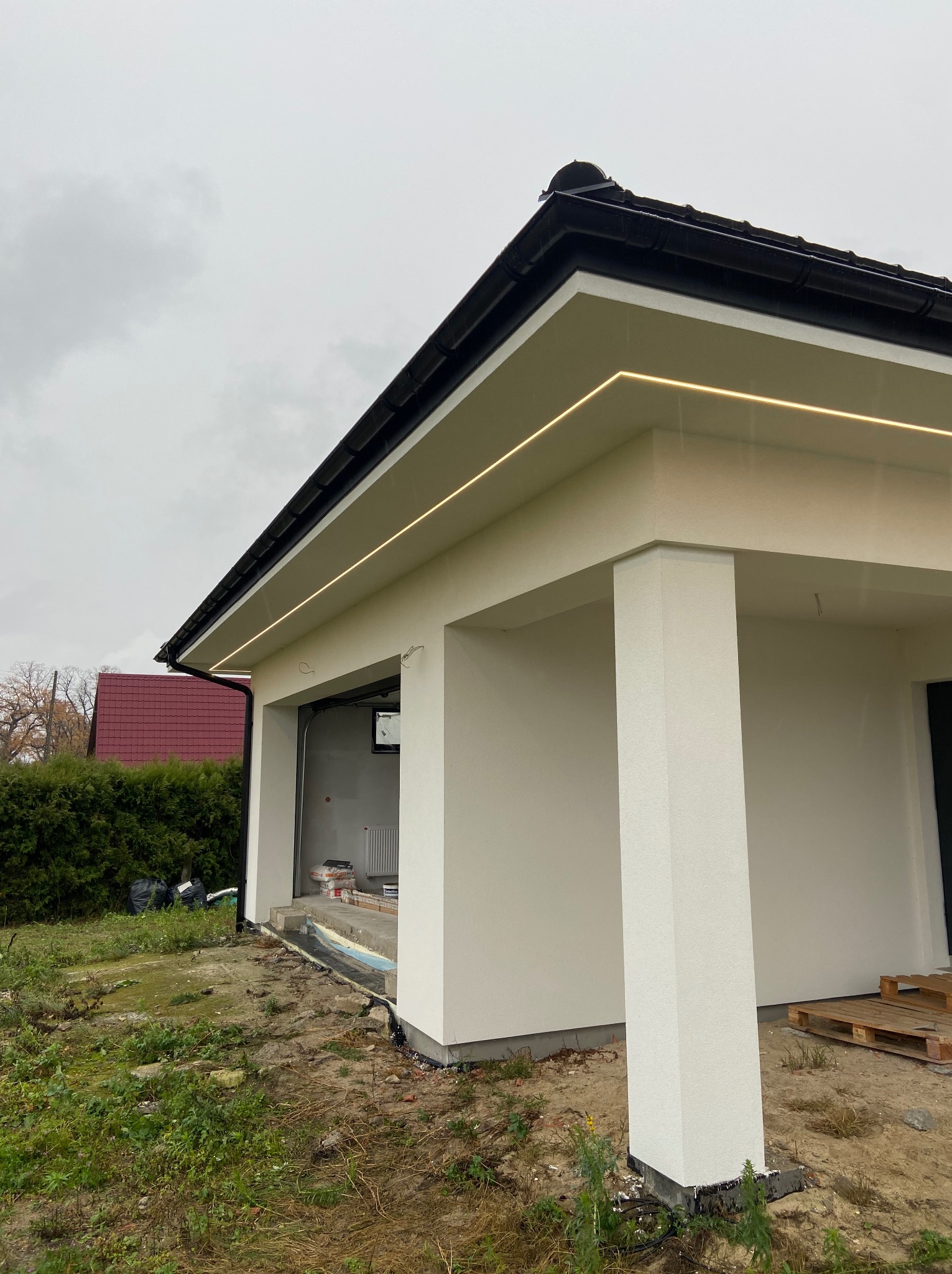 Nowoczesny dom z oświetleniem LED pod dachem, widok z zewnątrz. Biała elewacja, czarny dach, surowy stan otoczenia. Minimalistyczny design.