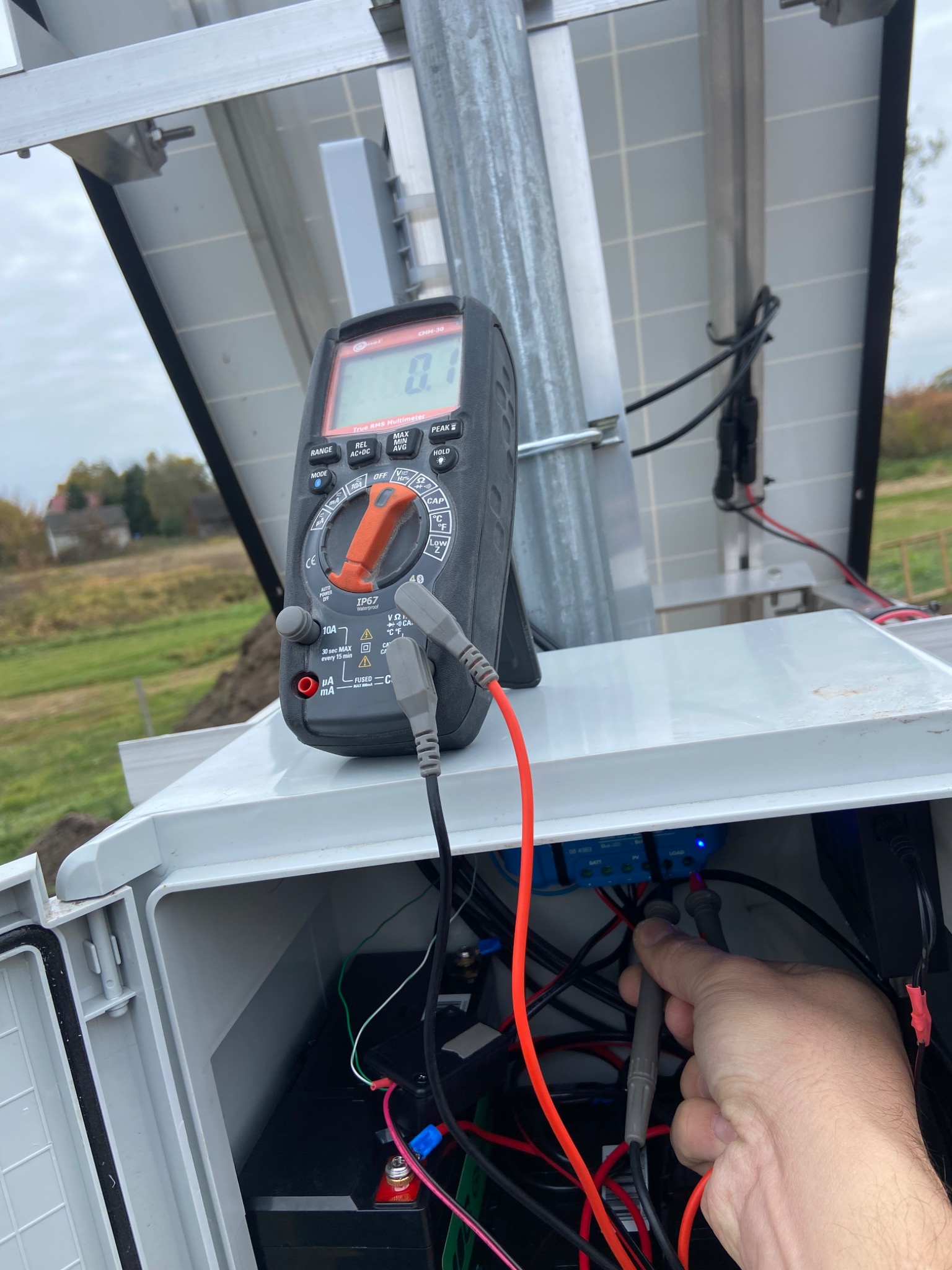 Pomiar napięcia instalacji fotowoltaicznej multimetrem. Widoczne panele, skrzynka z okablowaniem i akumulator. Praca w toku, pomiar parametrów.