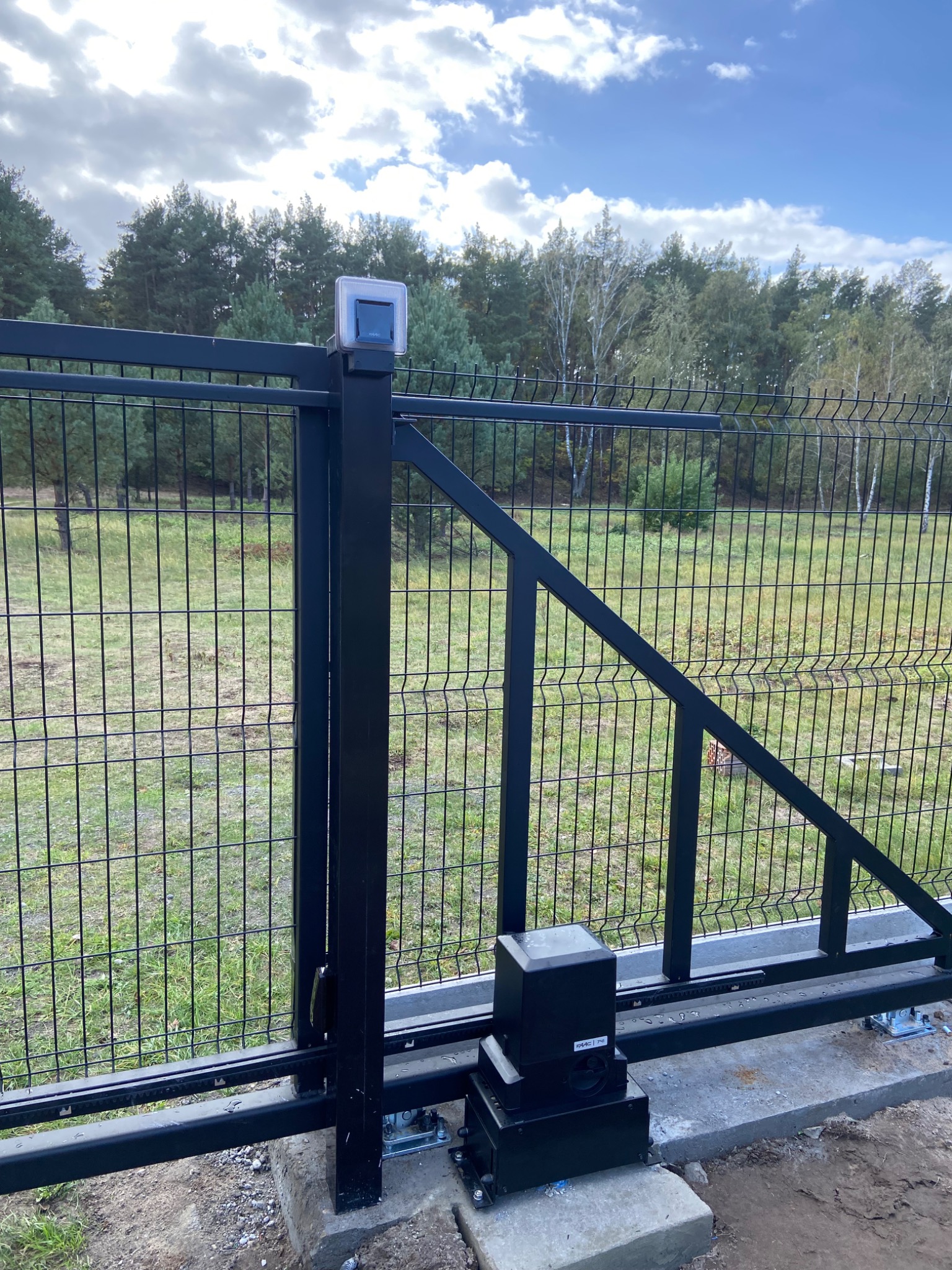 Czarna, metalowa brama przesuwna z automatyką, widoczna na tle zieleni i drzew. Brama z siatki, z napędem i lampą ostrzegawczą. Ujęcie z dołu, podkreślające mechanizm.