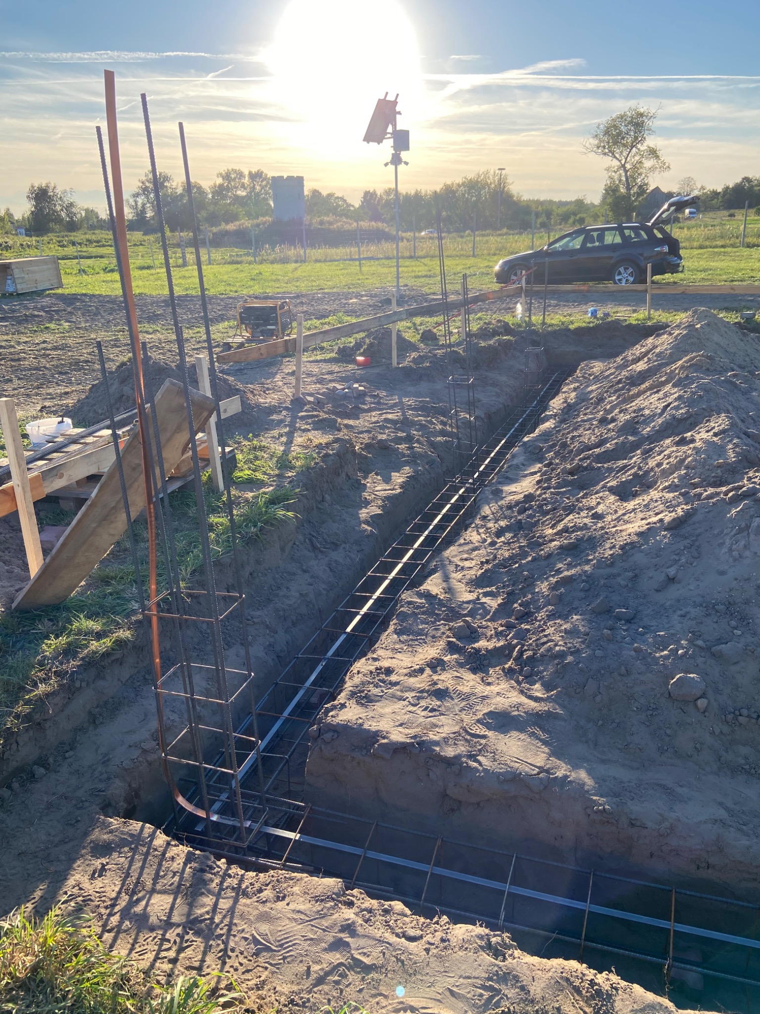 Wykonanie fundamentów: zbrojenie w wykopie, widoczne pręty i siatka stalowa. W tle krajobraz z samochodem i wieżą. Słoneczny dzień, budowa w toku.