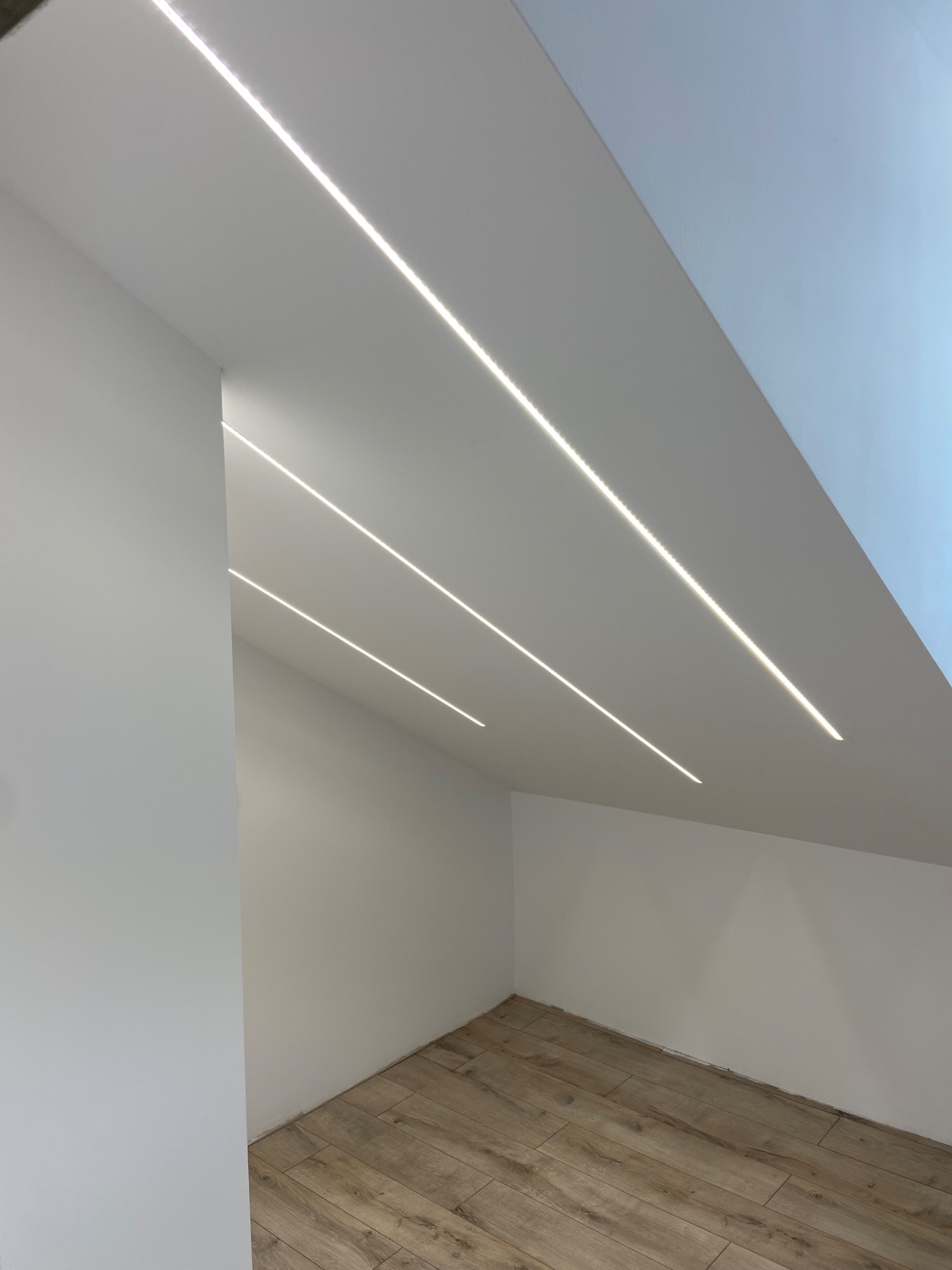 Nowoczesne oświetlenie LED w skosach poddasza, trzy równoległe linie światła, drewnopodobna podłoga, białe ściany. Minimalistyczny design.
