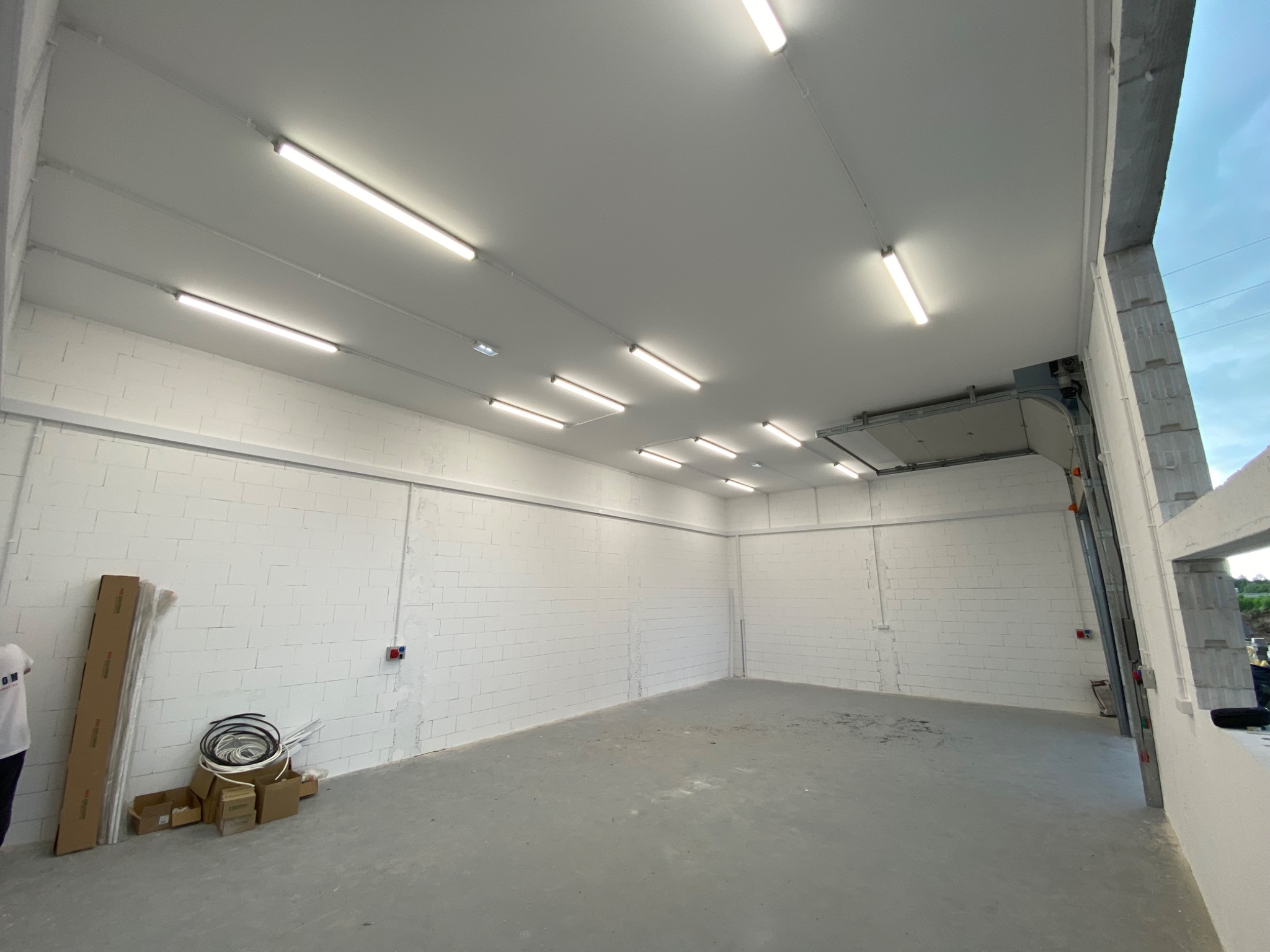 Wnętrze hali z białymi ścianami z betonu komórkowego i równomiernie rozmieszczonymi lampami LED. Widoczne kable i puszki elektryczne. Surowy beton na podłodze.
