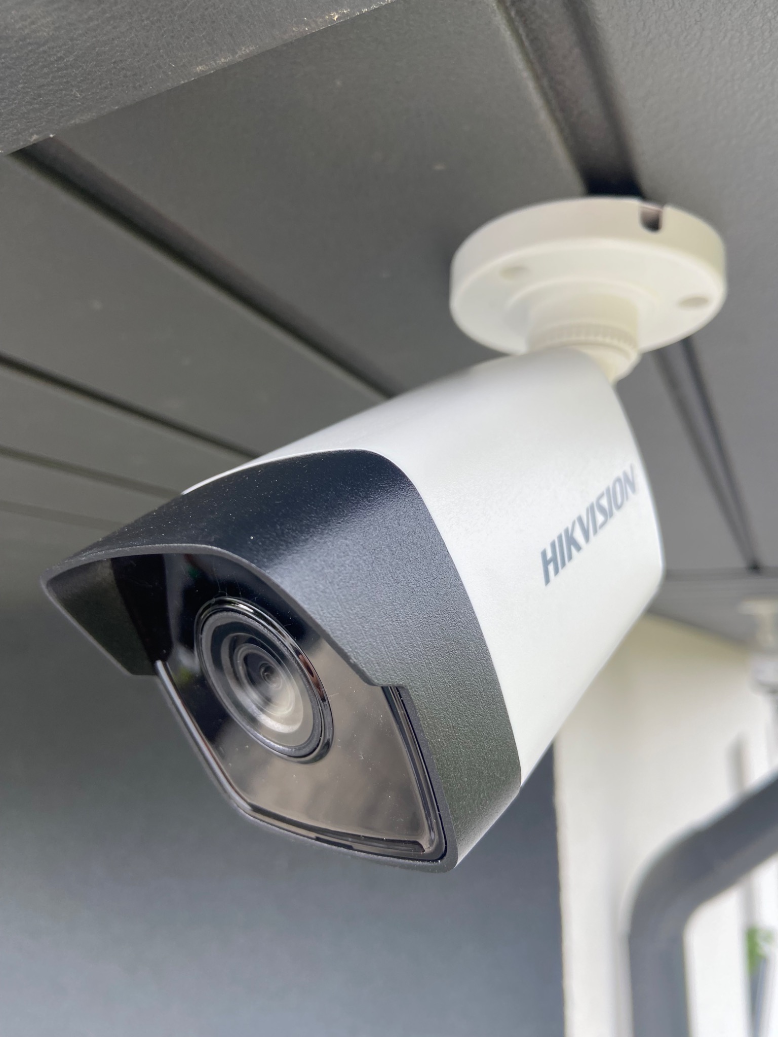 Zamontowana kamera monitoringu Hikvision, widoczna pod kątem, z czarnym daszkiem i szarym napisem na białej obudowie, przymocowana do szarego sufitu.