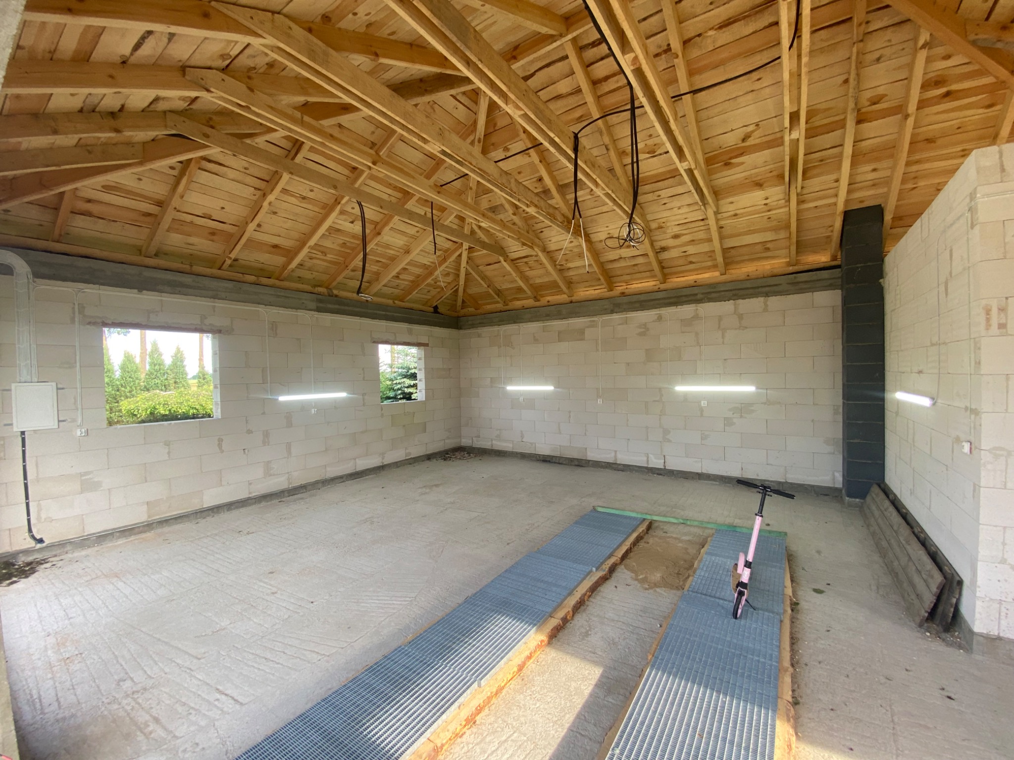 Wnętrze garażu z surowym drewnianym dachem, białymi ścianami z betonu komórkowego, oświetleniem LED i wystającymi kablami. Widoczne okna i metalowe kanały w podłodze.