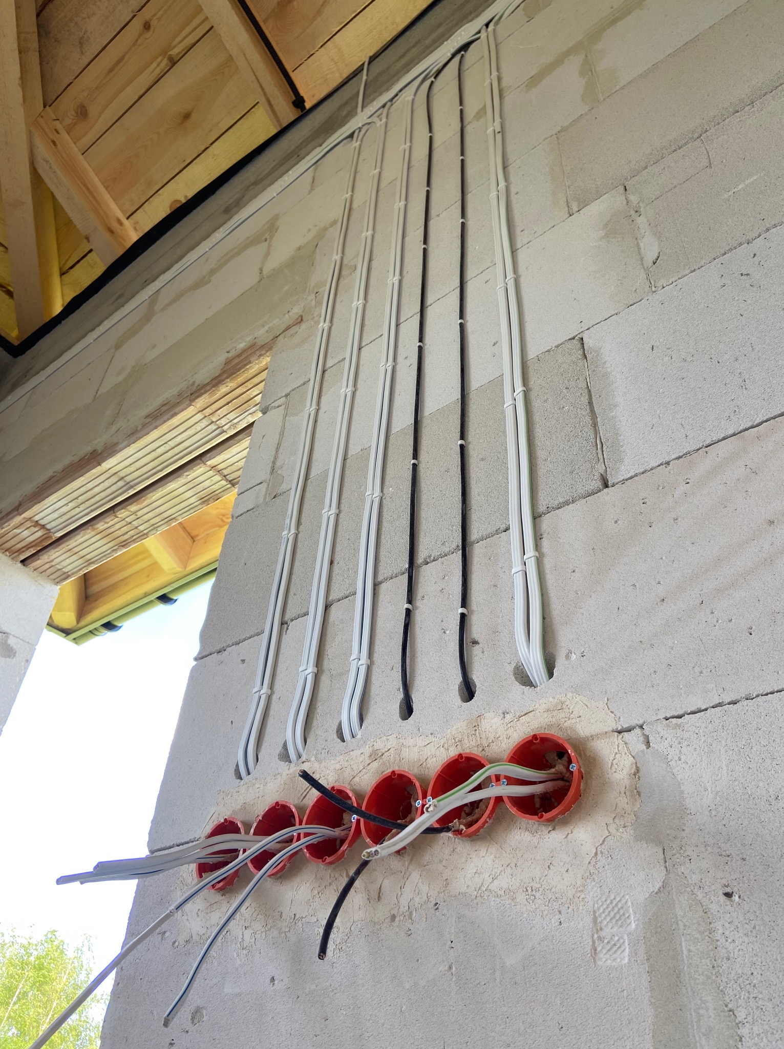 Instalacja elektryczna w ścianie z betonu komórkowego. Widoczne kable w peszlach i pomarańczowe puszki elektryczne. Częściowo widoczna konstrukcja drewniana dachu.