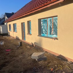 Bohdan Onyshchuk - Ściana domu po tynkowaniu, okna zabezpieczone folią. Widoczne detale: faktura tynku, dachówka, elementy zabezpieczające okna. Słoneczny dzień.