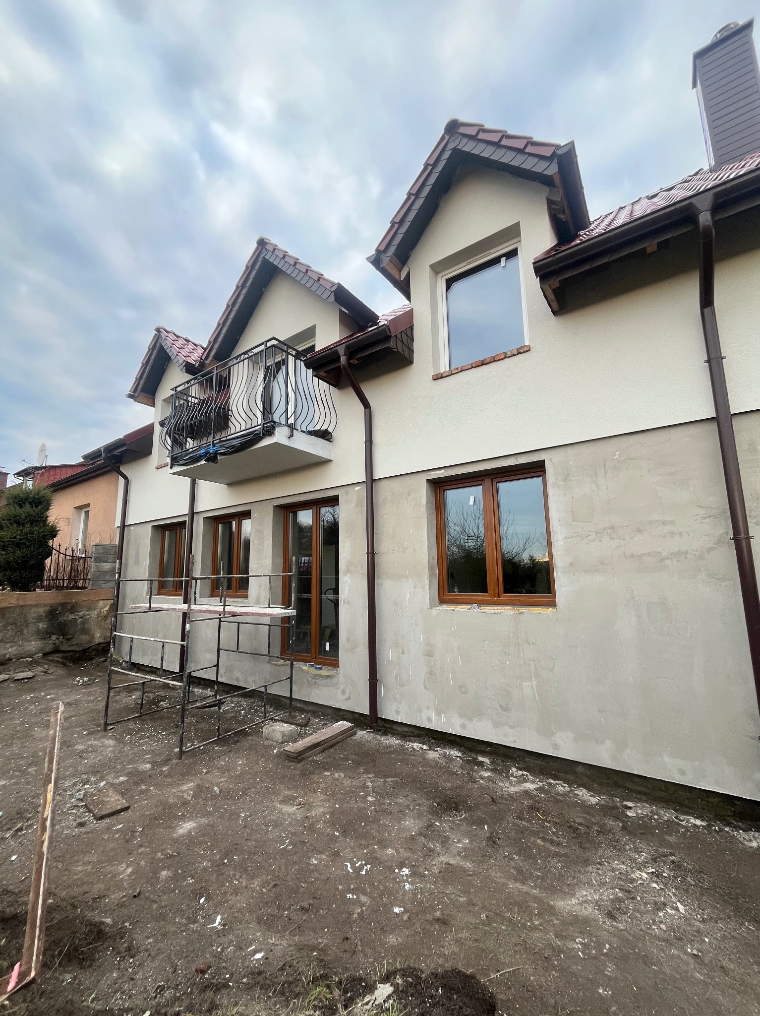 Dom z balkonem w trakcie prac elewacyjnych: górna część pomalowana na kremowo, dolna w szarym tynku. Widoczne rusztowania i brązowe rynny.