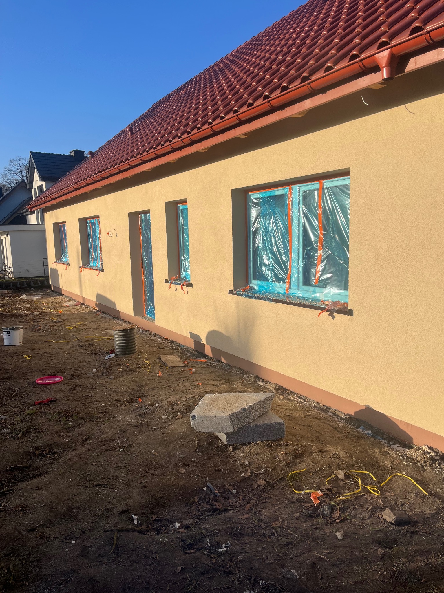 Ściana domu po tynkowaniu, okna zabezpieczone folią. Widoczne detale: faktura tynku, dachówka, elementy zabezpieczające okna. Słoneczny dzień.