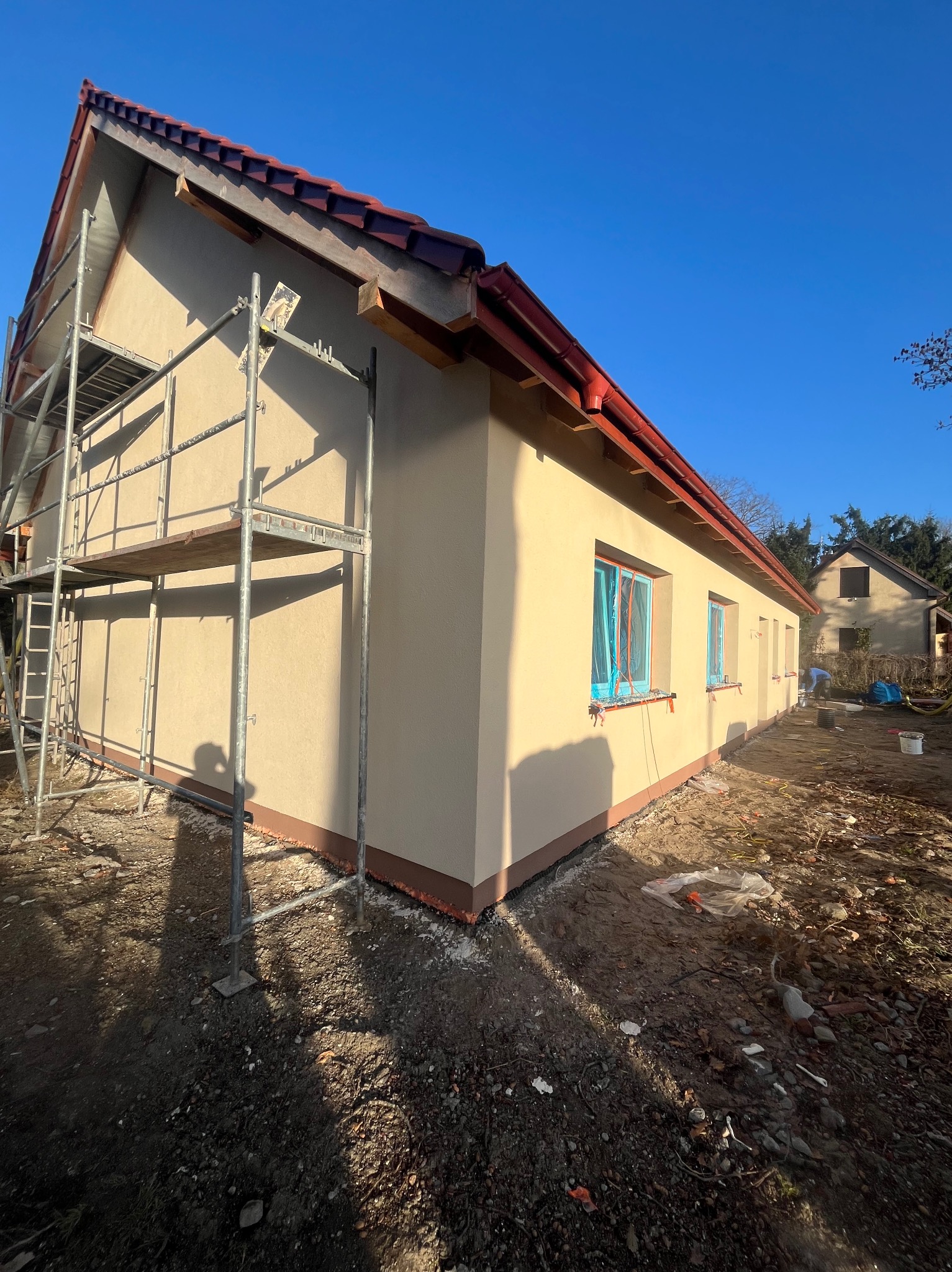 Budynek po tynkowaniu z rusztowaniem. Okna zabezpieczone folią. Widoczne detale wykończenia i przygotowania do dalszych prac elewacyjnych w słoneczny dzień.