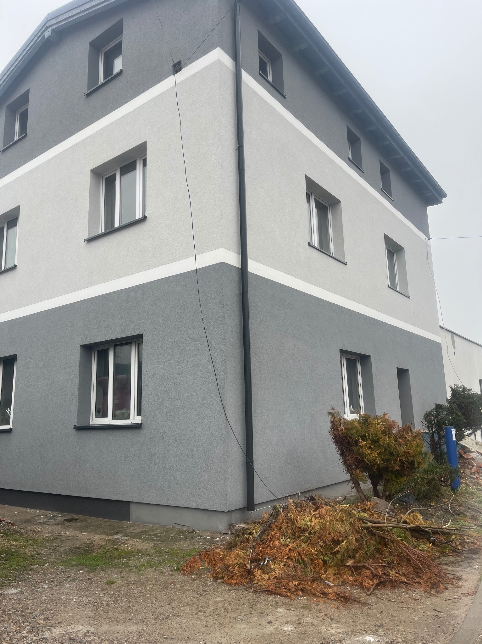 Dwukolorowa elewacja budynku z wyraźnym podziałem na szary i jasnoszary pas, widok z dołu, zieleń u podstawy. Nowoczesne wykończenie.