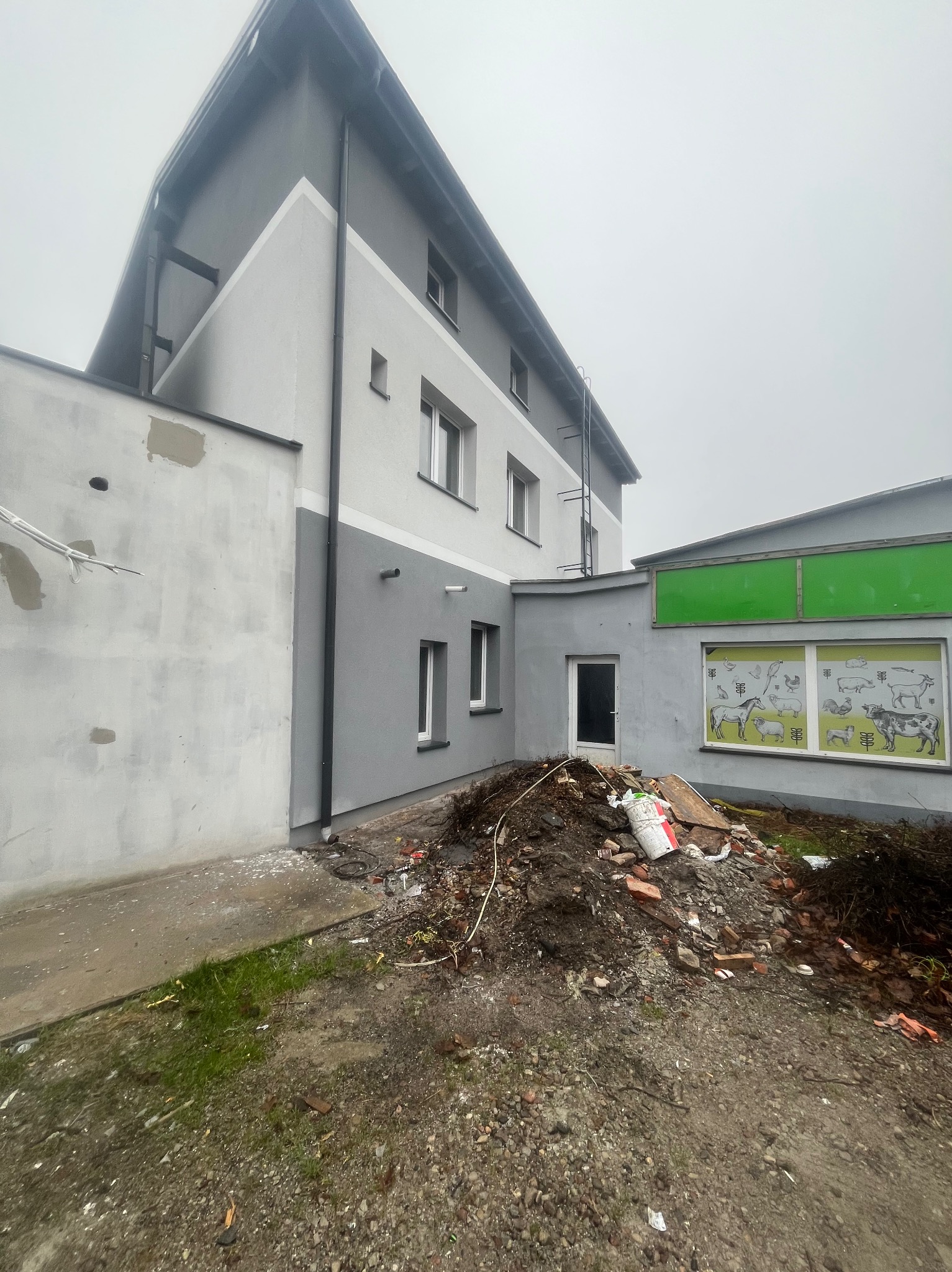 Budynek po częściowej renowacji elewacji, z widocznymi śladami prac i gruzem. Fragment budynku z nowym szarym tynkiem kontrastuje z surową, białą ścianą.