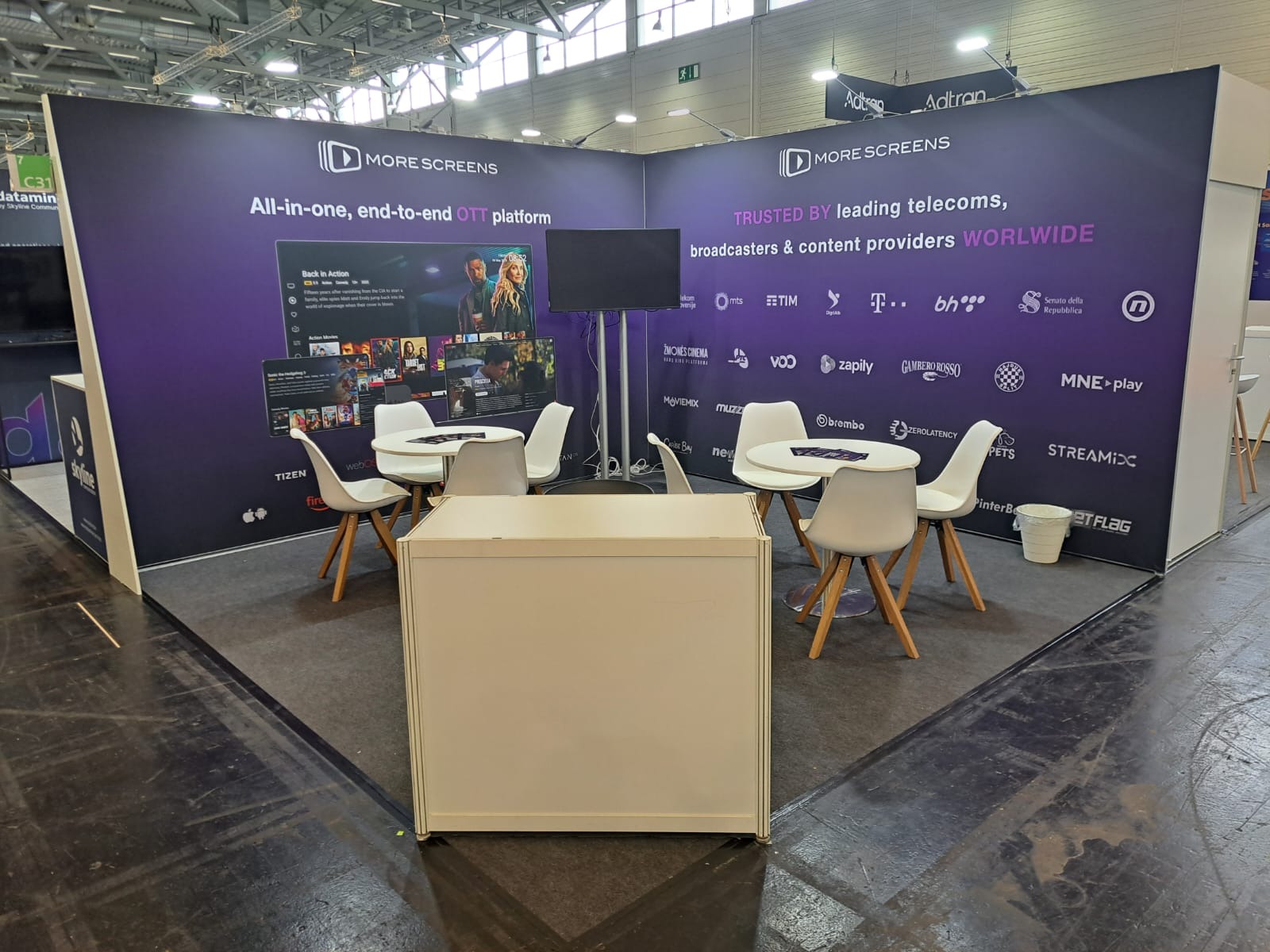 Stoisko targowe More Screens z fioletowym tłem, białymi krzesłami i stolikami. Widoczne logo firmy, ekran i lista partnerów. Oświetlenie sufitowe i ciemna podłoga.