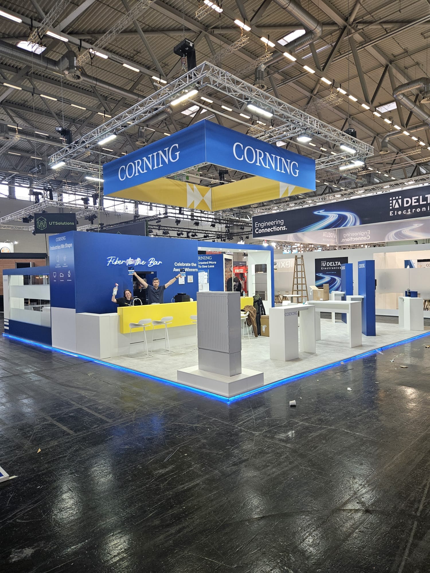 Stoisko targowe Corning z podświetleniem LED, prezentujące produkty i usługi. Widoczni pracownicy za ladą, banery reklamowe i elementy ekspozycji.