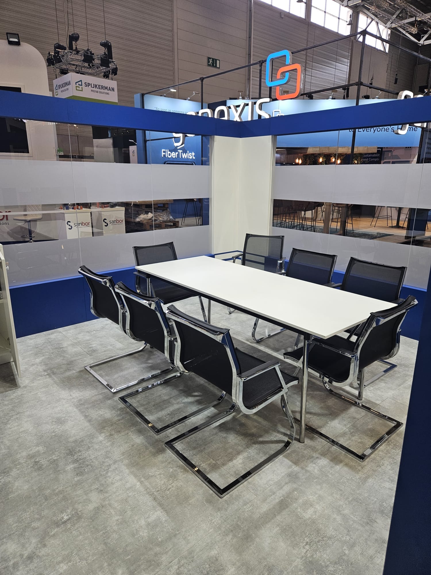 Nowoczesna sala konferencyjna z białym stołem i czarnymi krzesłami na szarym tle. Widoczne logo firmy i elementy wystawiennicze.