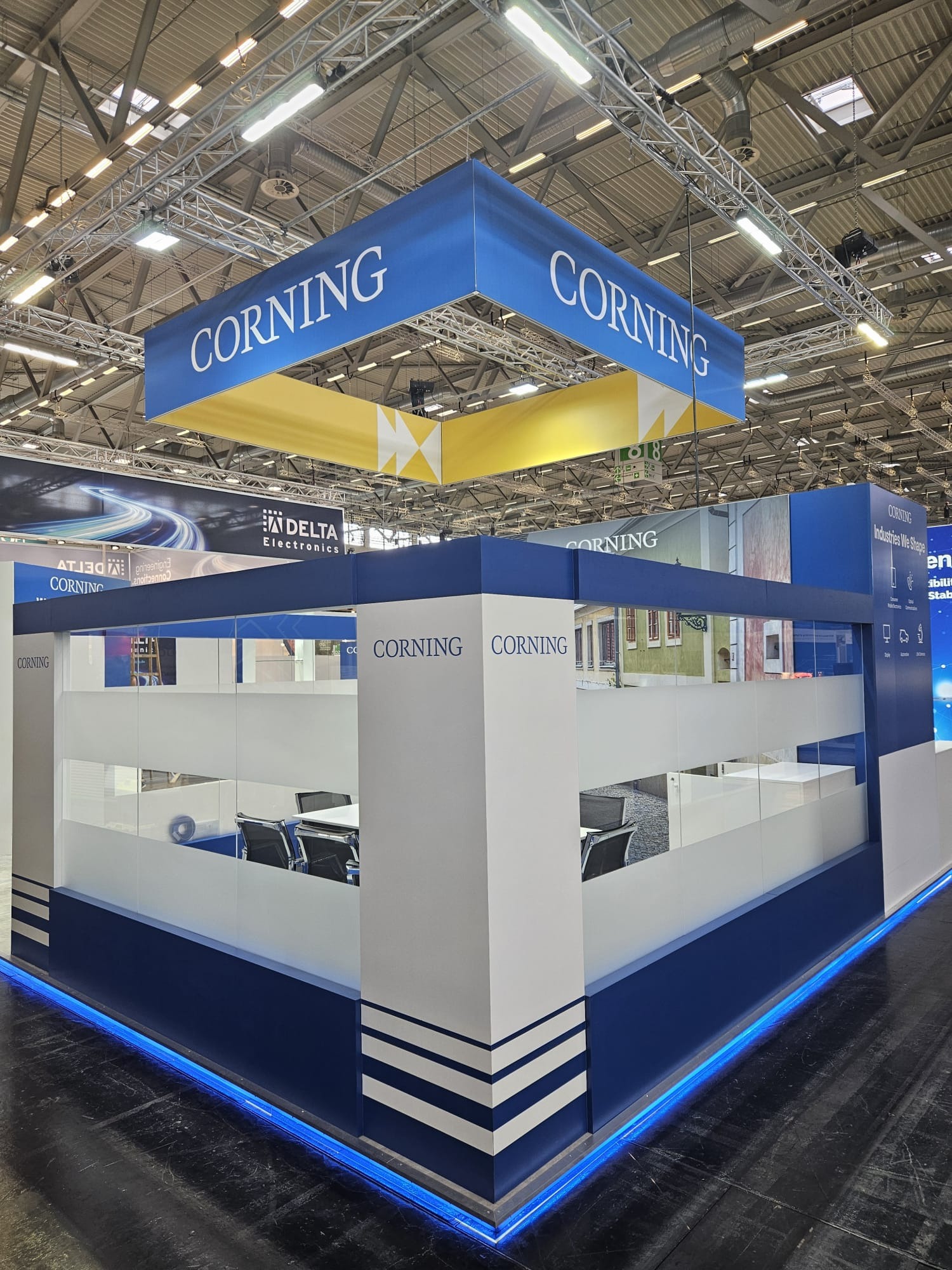 Stoisko targowe Corning z niebieskim oświetleniem LED, logo na górze i ścianach. Wewnątrz widoczne krzesła i stół. Nowoczesny design, jasne kolory, widok z perspektywy.