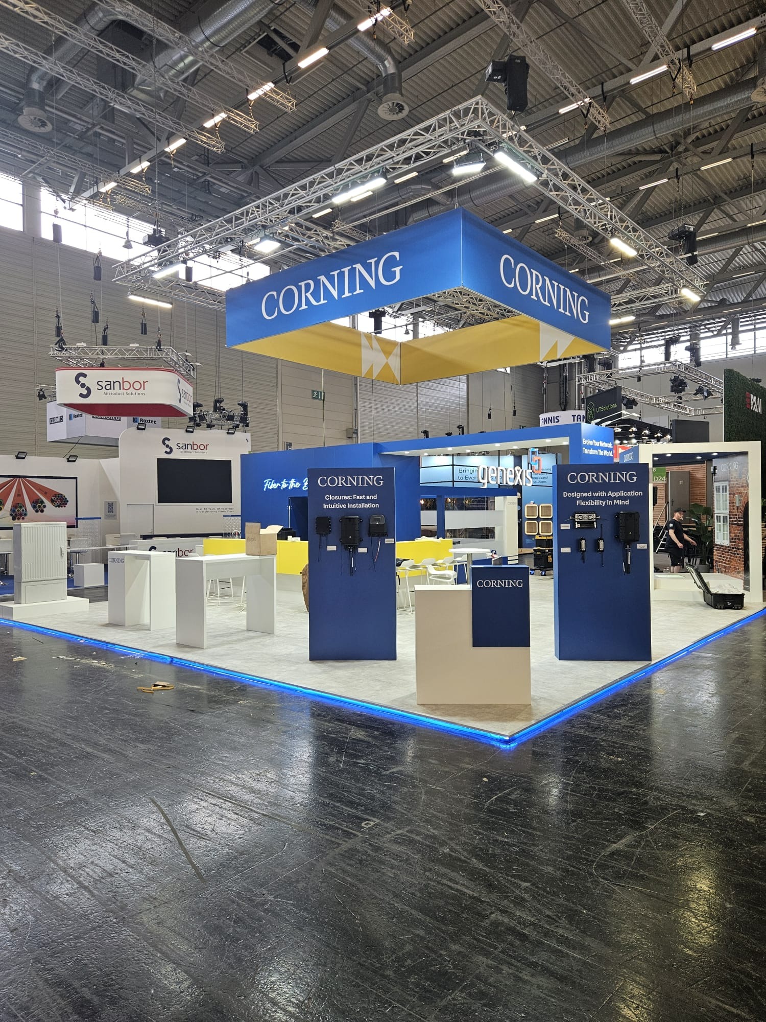Stoisko targowe Corning z niebieskimi akcentami świetlnymi, prezentujące produkty i rozwiązania telekomunikacyjne. Widoczne logo i hasła reklamowe.