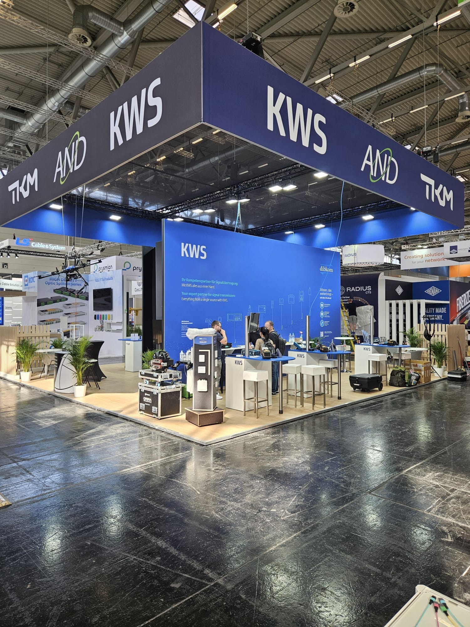 Stoisko targowe KWS z niebieskimi ścianami, białymi meblami i podwieszonym banerem z logo. Widoczni ludzie przy stanowiskach, prezentacja produktów i oświetlenie.