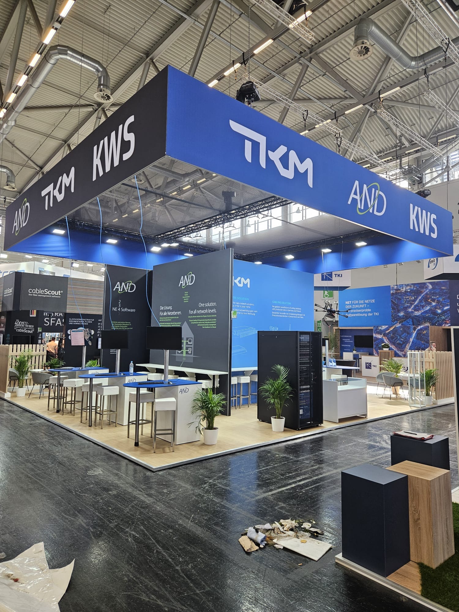 Stoisko targowe z logo firm TKM i AND KWS. Widoczne meble, rośliny doniczkowe i sprzęt IT. Podwieszana konstrukcja z brandingiem. Na podłodze widoczne ślady po montażu.