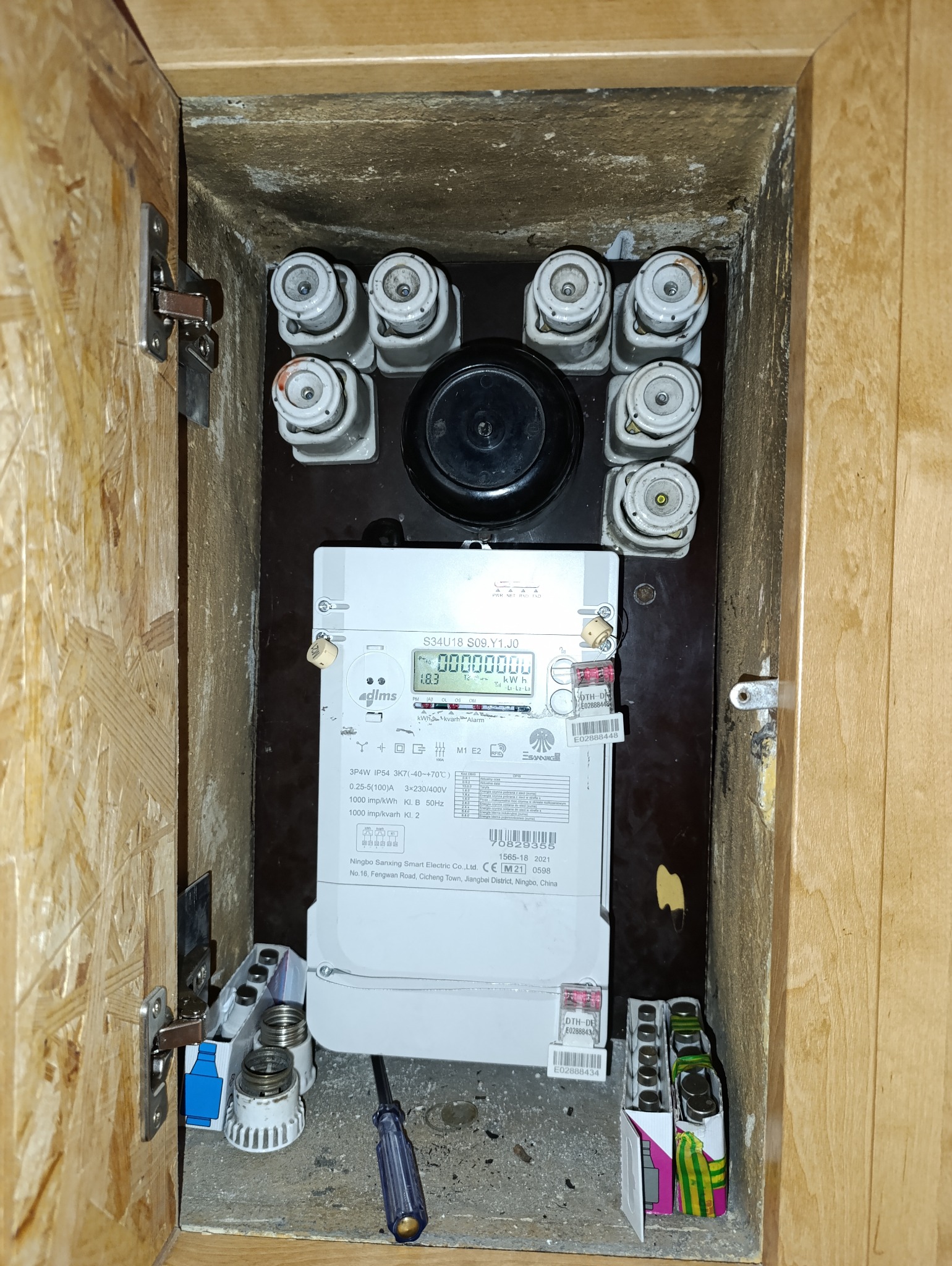 Otwarta skrzynka z licznikiem elektrycznym, bezpiecznikami topikowymi i zapasowymi wkładkami. Widoczny śrubokręt. Ujęcie z bliska, wnętrze szafki z płyty OSB.