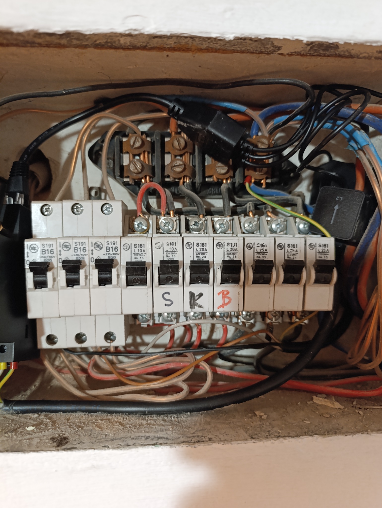 Otwarta skrzynka rozdzielcza z bezpiecznikami i przewodami. Widoczne oznaczenia na bezpiecznikach i ręczne dopiski 'SKB'. Ujęcie z bliska, ukazujące detale instalacji.