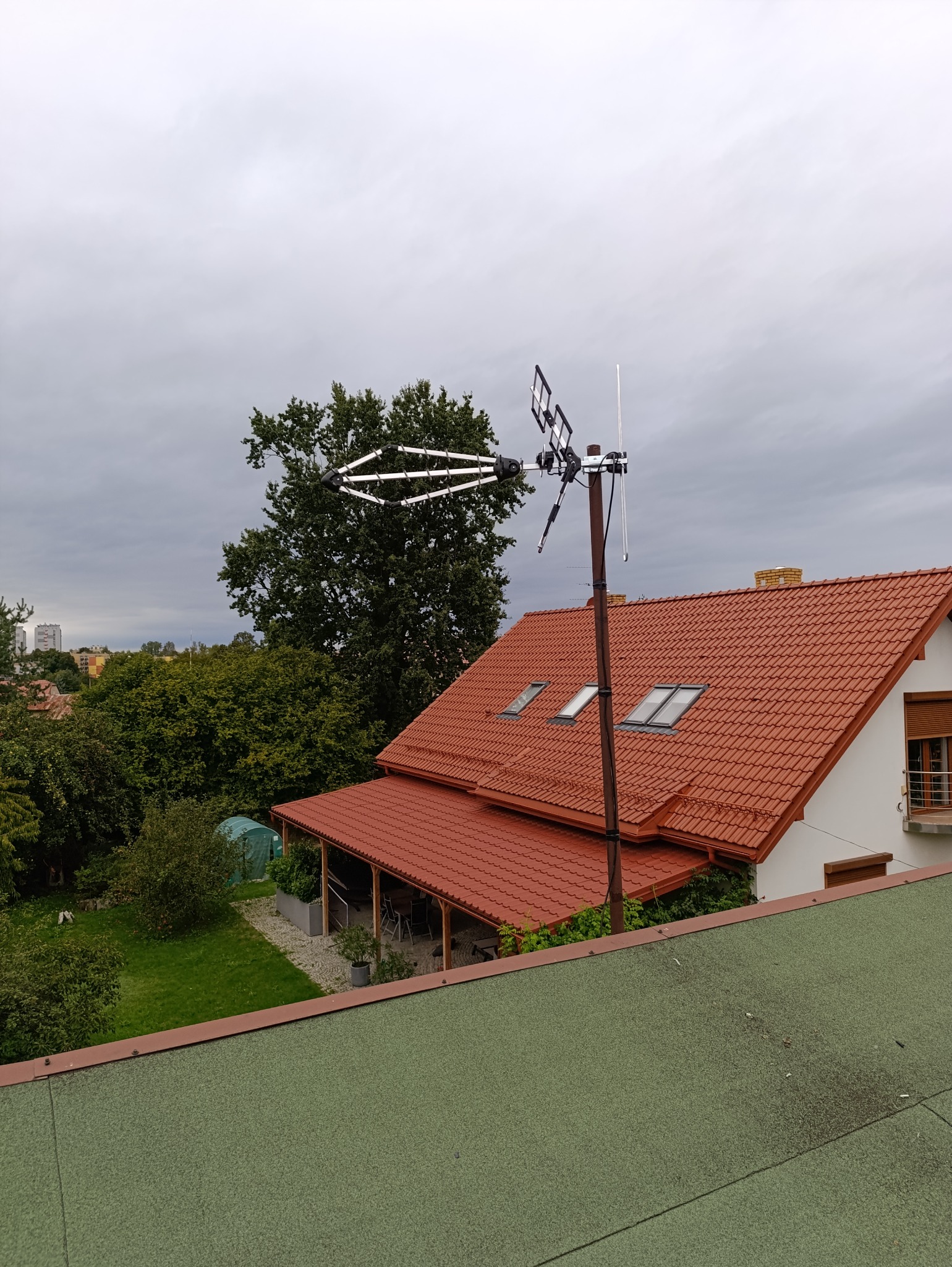 Antena telewizyjna zamontowana na dachu domu jednorodzinnego z czerwoną dachówką, widok z góry na tle zieleni i pochmurnego nieba.