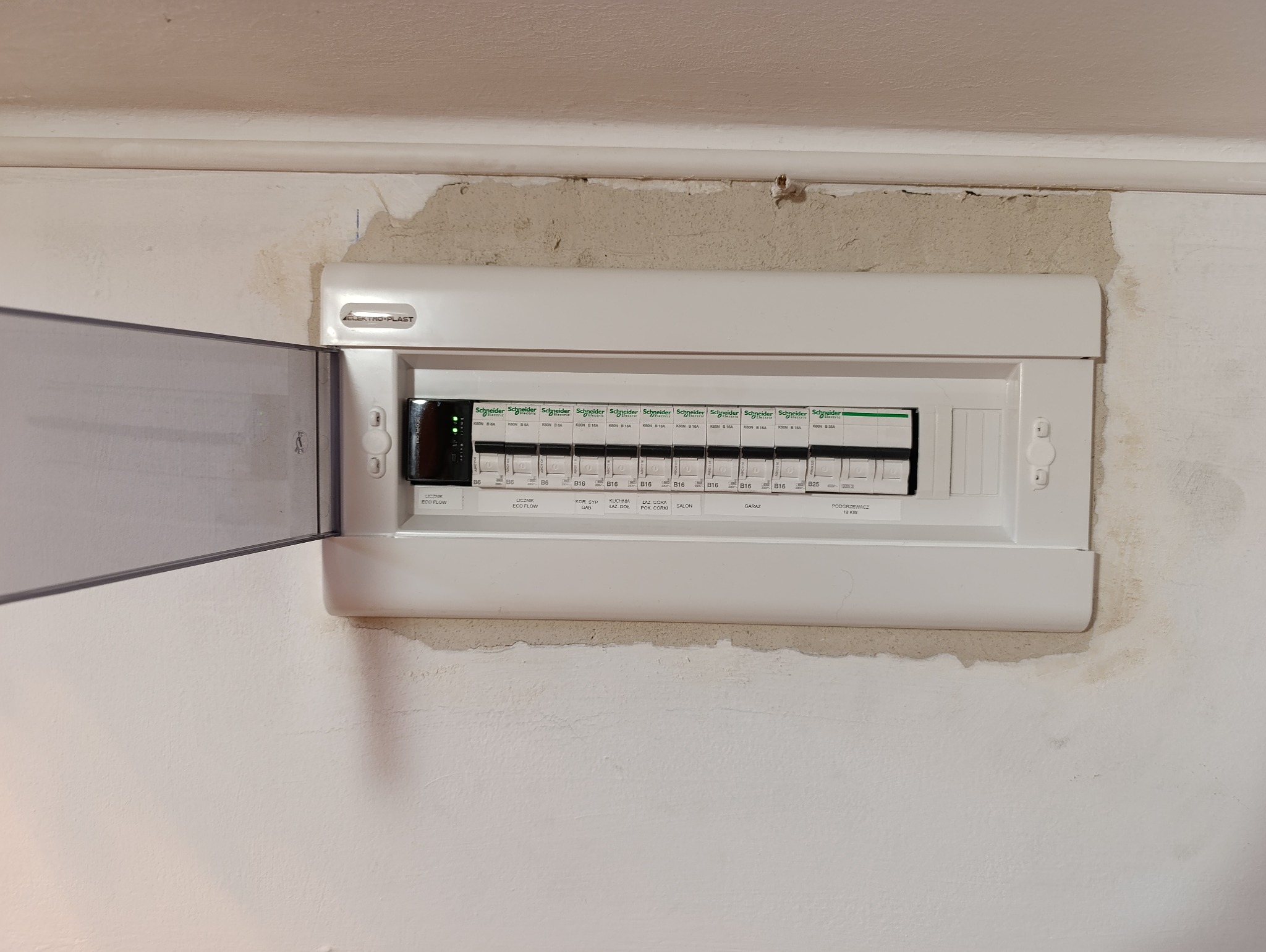Rozdzielnia elektryczna Schneider z otwartą osłoną, zamontowana na ścianie z widocznymi śladami naprawy. Widoczne bezpieczniki i oznaczenia obwodów.