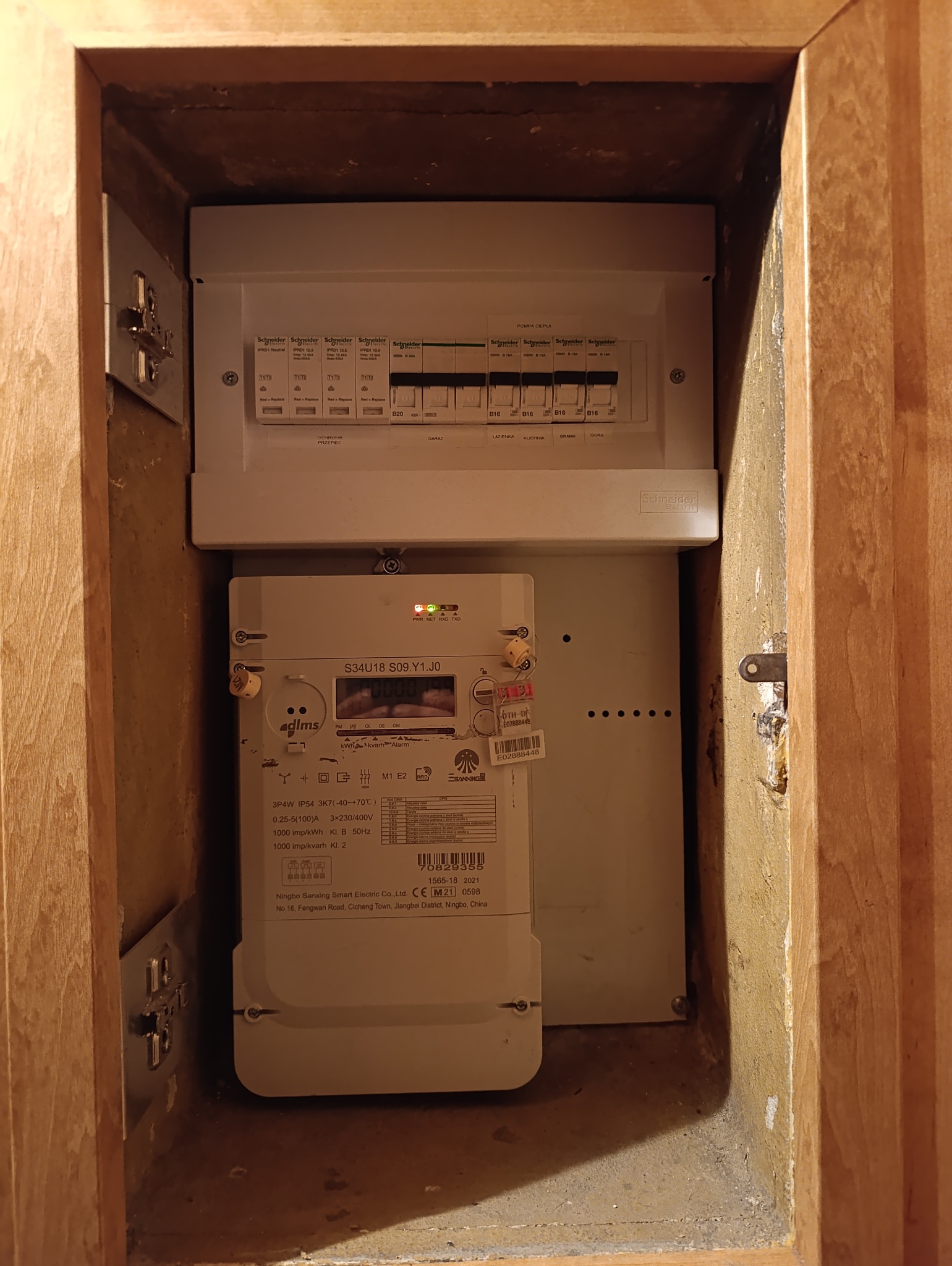 Rozdzielnia elektryczna po modernizacji w drewnianej obudowie. Widoczny licznik energii i zabezpieczenia Schneider Electric. Oświetlenie górne.