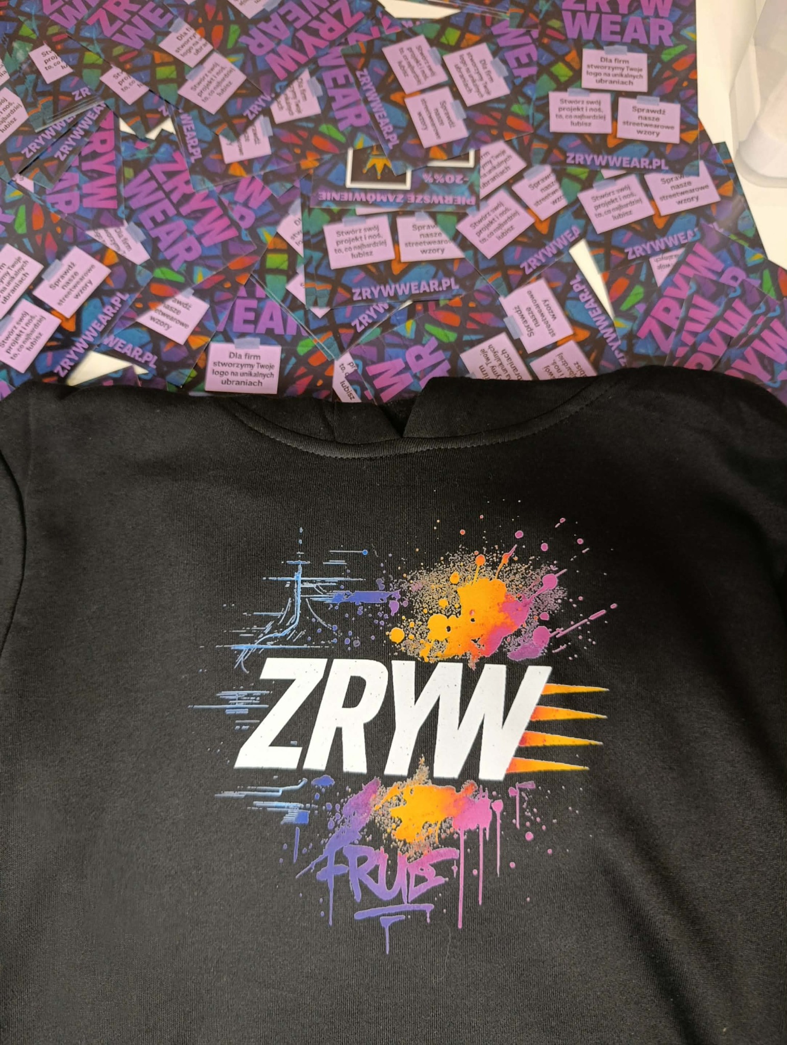 Czarna bluza z nadrukiem 'ZRYW' i kolorowymi plamami, otoczona ulotkami z logo firmy ZRYWWEAR.PL, oferującej tworzenie logo na odzieży. Streetwearowe wzory.