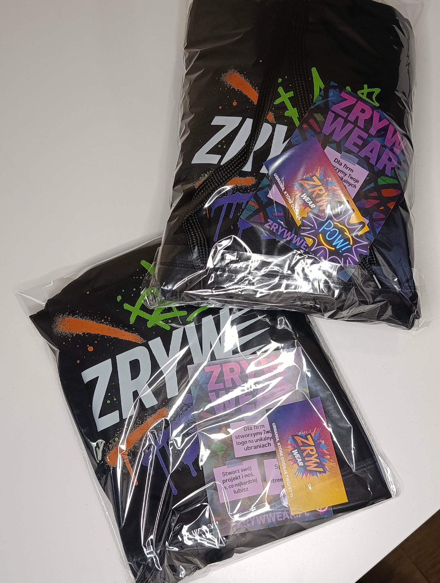 Dwa zapakowane czarne T-shirty z kolorowym nadrukiem 'ZRYW WEAR' i ulotkami informacyjnymi, leżące na białym tle. Widoczne detale graficzne i logo marki.
