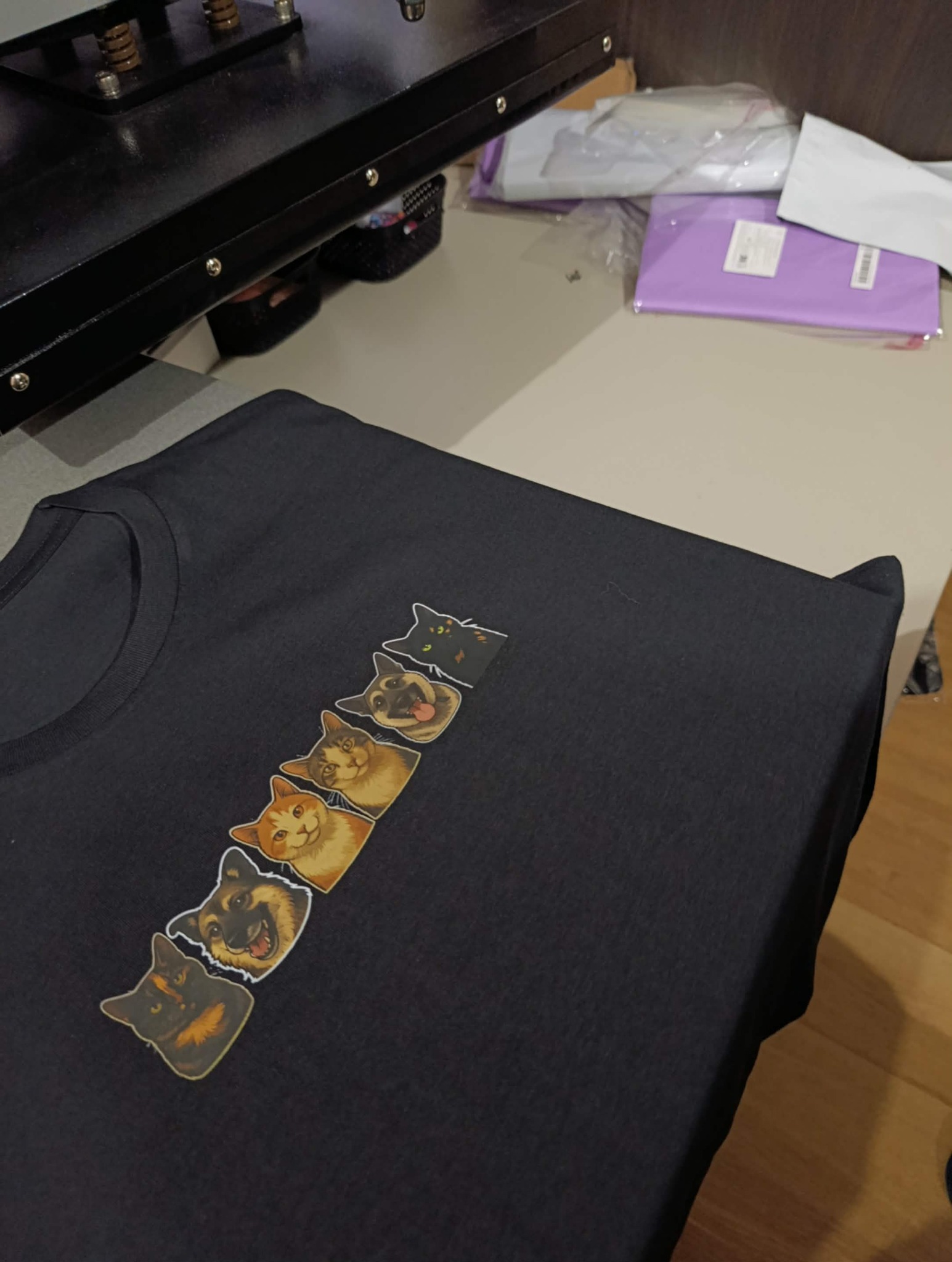 Czarna koszulka z rzędem naklejonych, zabawnych portretów zwierząt (koty, pies) pod prasą termotransferową. Gotowy produkt po nadruku.