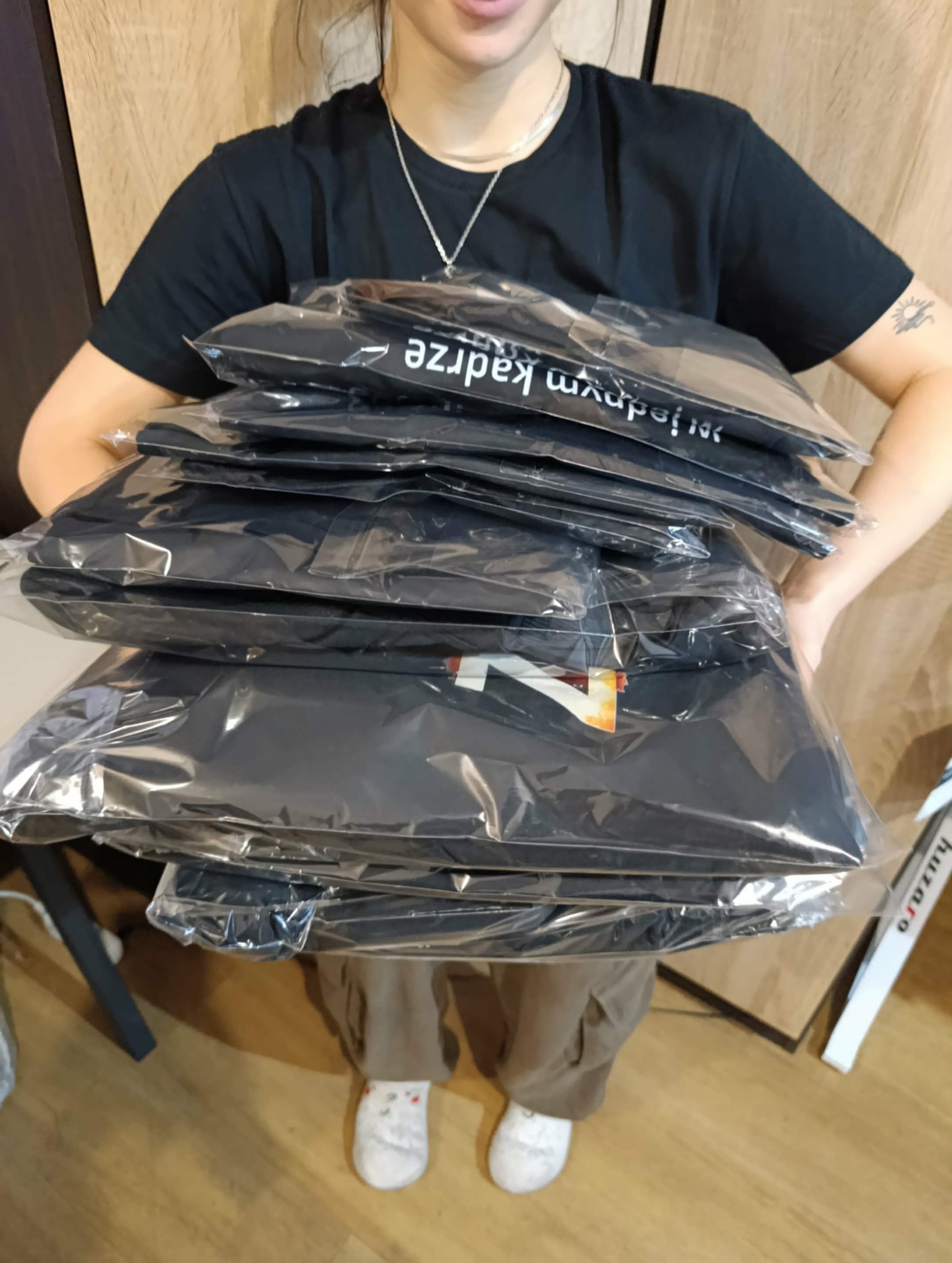 Osoba trzyma stertę zapakowanych koszulek z nadrukami, widoczne logo '7' i napis 'i jednym kadrem'.