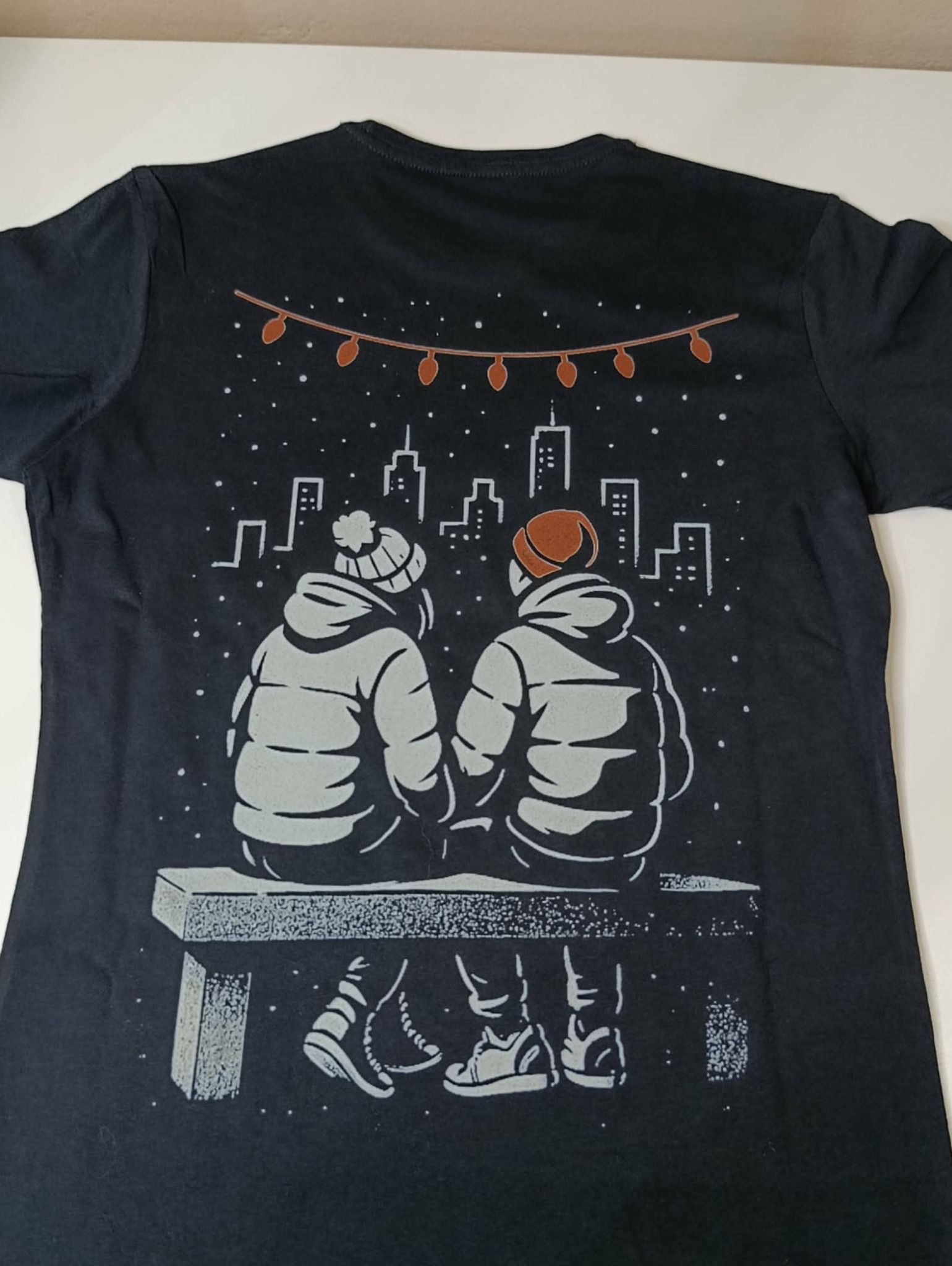 Czarna koszulka z nadrukiem przedstawiającym parę siedzącą na ławce na tle panoramy miasta i girlandy światełek. Grafika w odcieniach szarości i brązu.