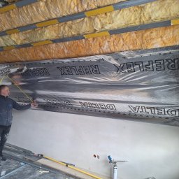 Kamil Stępiński - Pracownik mierzy folię refleksyjną na suficie z izolacją z wełny mineralnej. Widoczne profile metalowe i narzędzia. Etap prac wykończeniowych wewnątrz budynku.