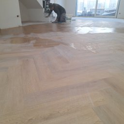Kamil Stępiński - Parkiet w jodełkę zabezpieczany lakierem przez pracownika. Widoczne wiadro z lakierem i narzędzia. Jasne wnętrze z dużym oknem. Etap prac wykończeniowych.