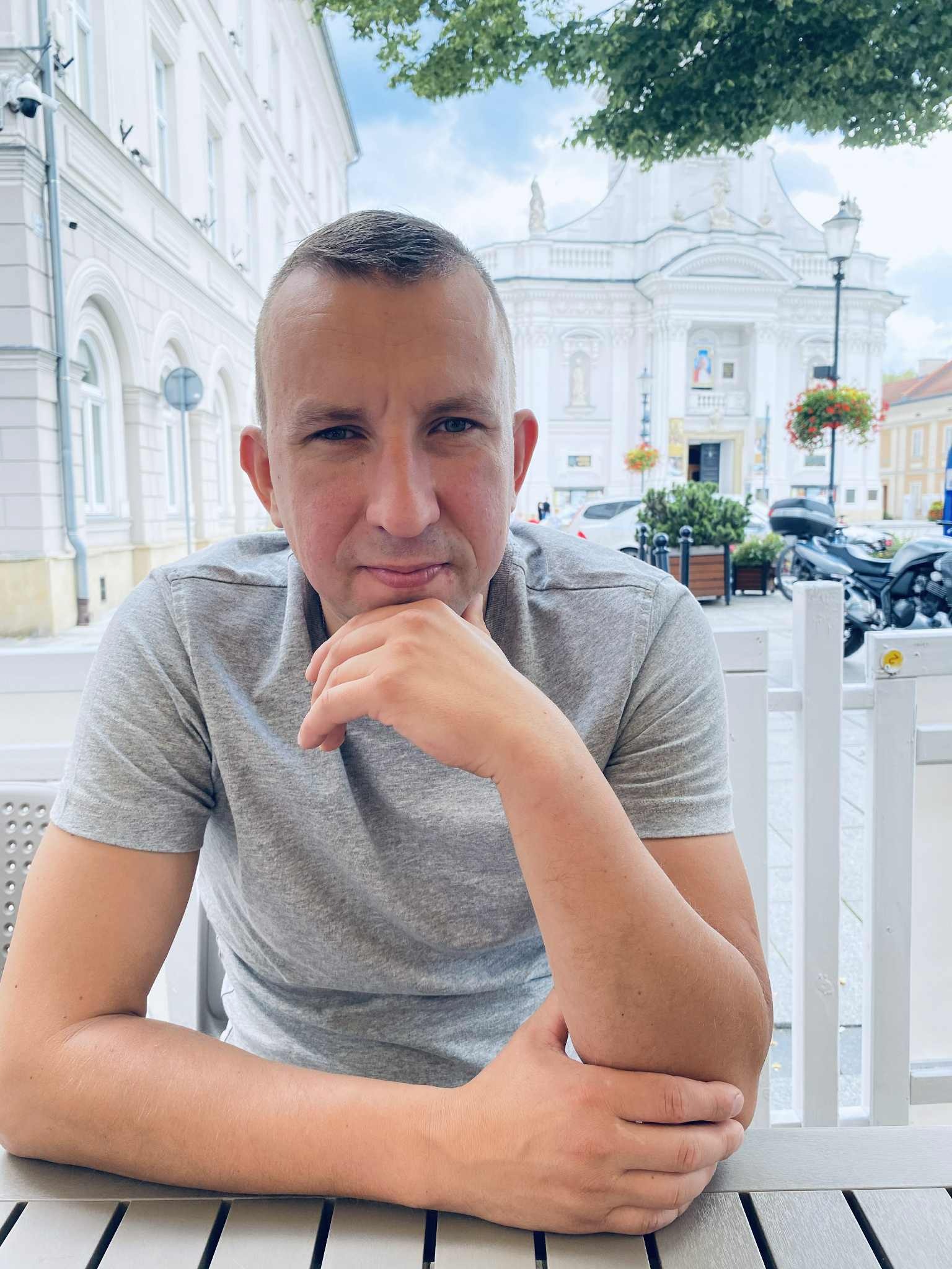 Portret mężczyzny w szarej koszulce na tle architektury miejskiej Chorzowa. Widoczny budynek z kolumnami i dekoracjami, motocykle i zieleń w tle.