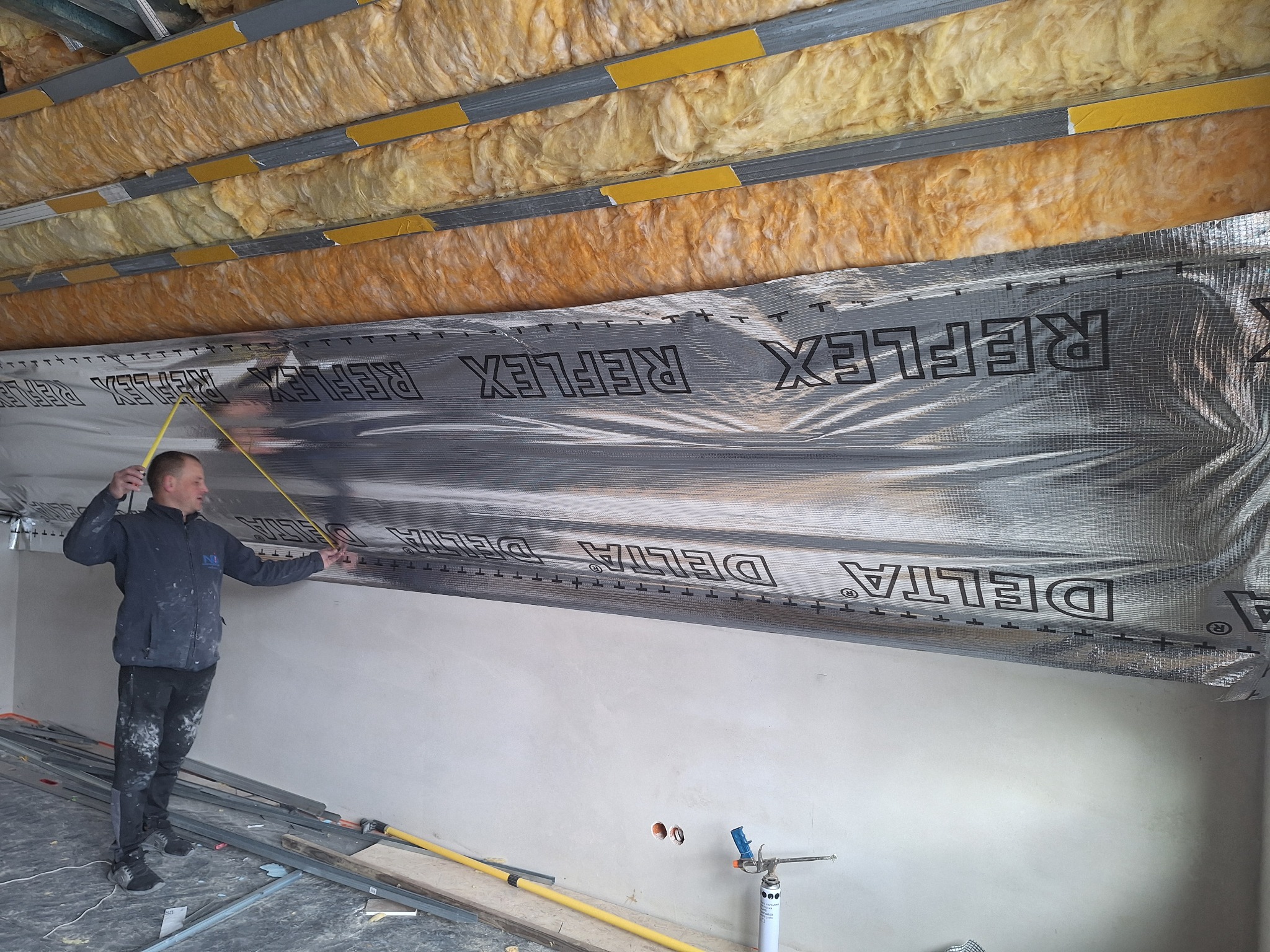 Pracownik mierzy folię refleksyjną na suficie z izolacją z wełny mineralnej. Widoczne profile metalowe i narzędzia. Etap prac wykończeniowych wewnątrz budynku.
