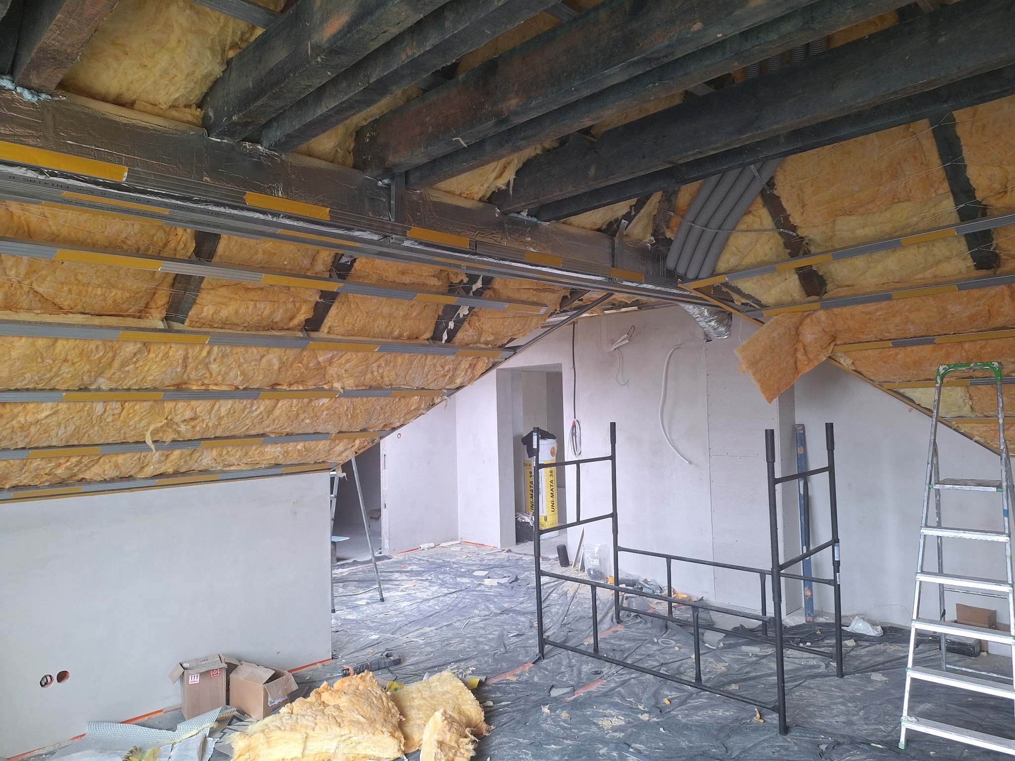 Poddasze w trakcie izolacji: wełna mineralna między krokwiami, metalowe profile, folia paroizolacyjna. Widoczne narzędzia i materiały budowlane. Etap prac wykończeniowych.