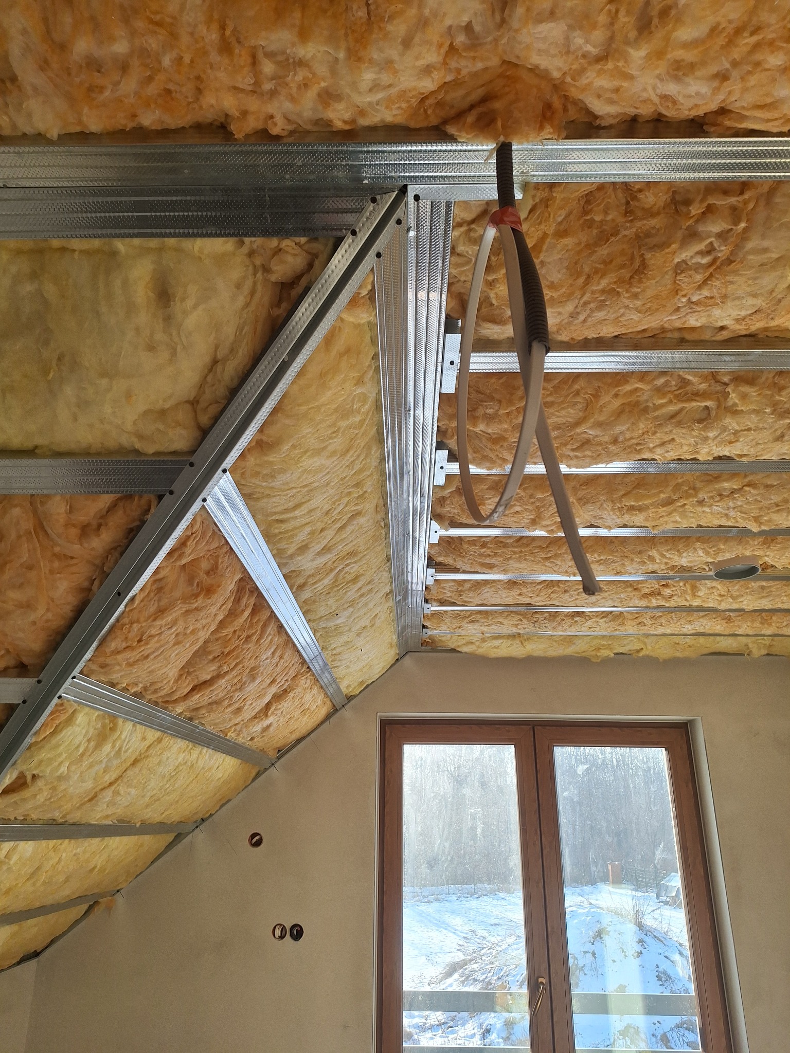 Skośny sufit poddasza z izolacją z wełny mineralnej, widoczne metalowe profile konstrukcyjne i okno dachowe z widokiem na zimowy krajobraz. Prace wykończeniowe.