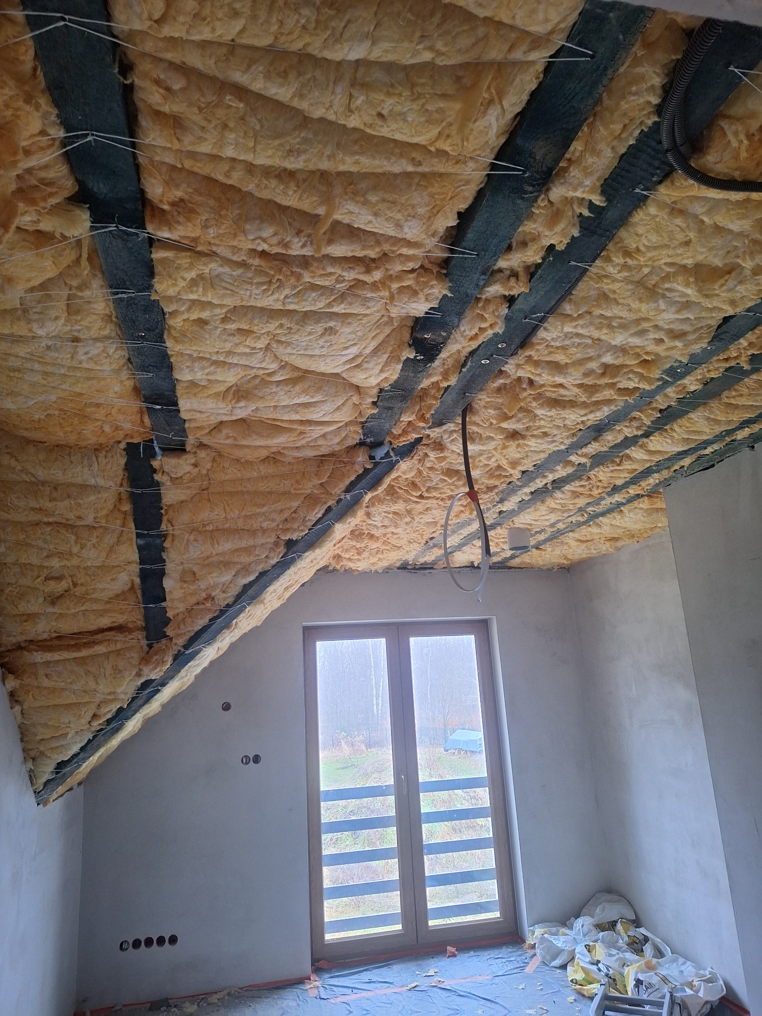 Ocieplenie poddasza wełną mineralną między krokwiami, zabezpieczone sznurkiem, widok z pokoju w Chorzowie. Okno dachowe w tle.