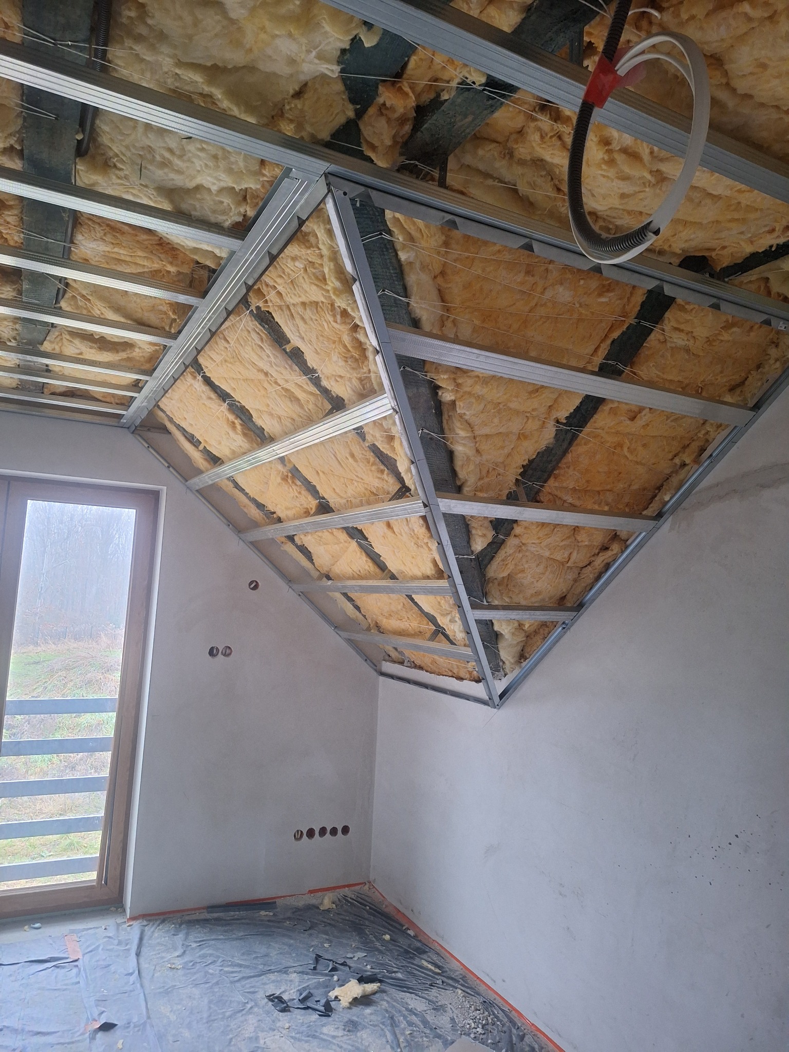 Sufit podwieszany z izolacją z wełny mineralnej w trakcie montażu. Widoczne metalowe profile i instalacja elektryczna. Okno w tle. Podłoga zabezpieczona folią.