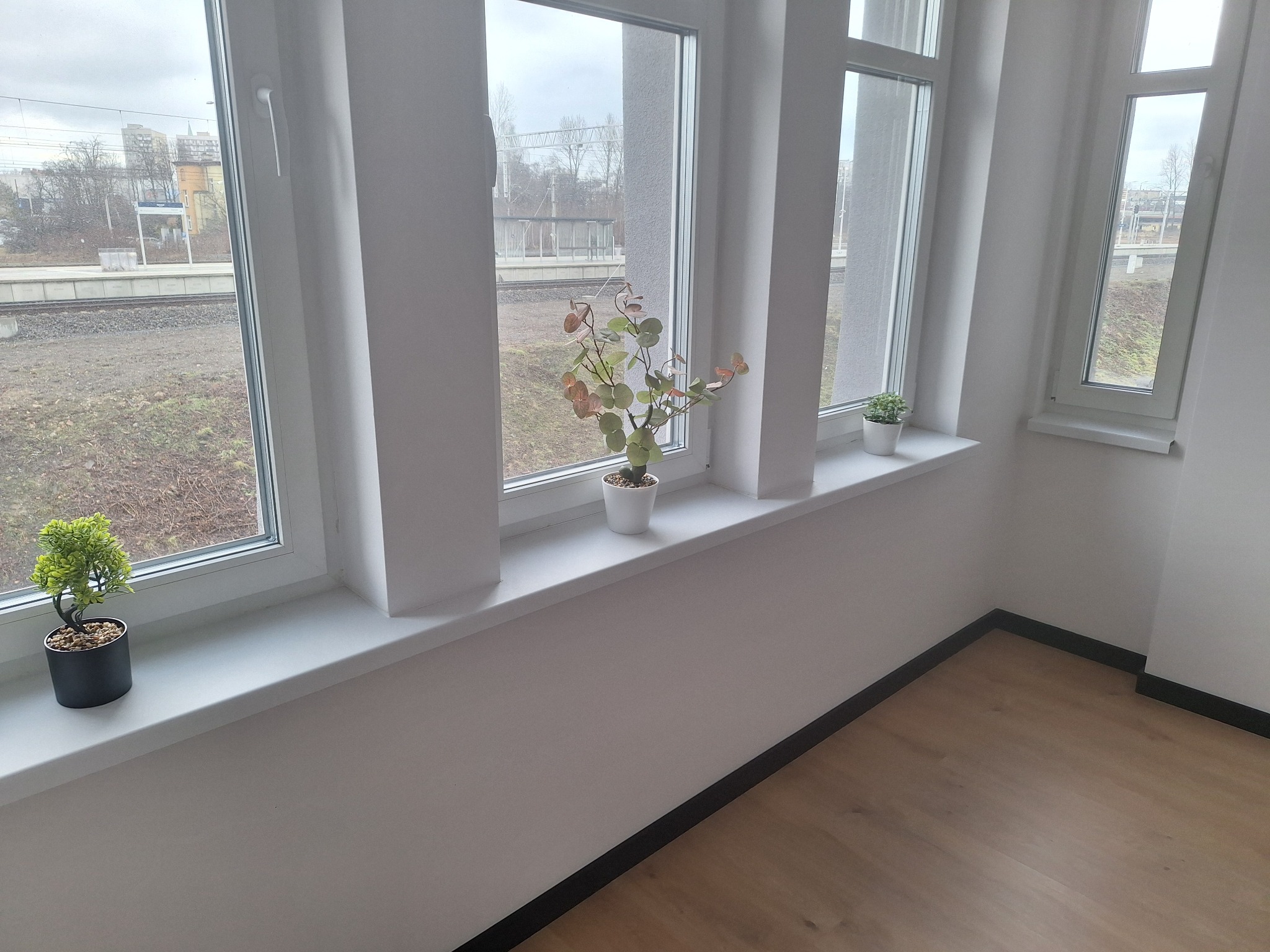 Jasne wnętrze z trzema oknami PCV, na których stoją doniczki z roślinami. Widok na zewnątrz ukazuje tory kolejowe i budynki w oddali. Podłoga wykończona panelami.
