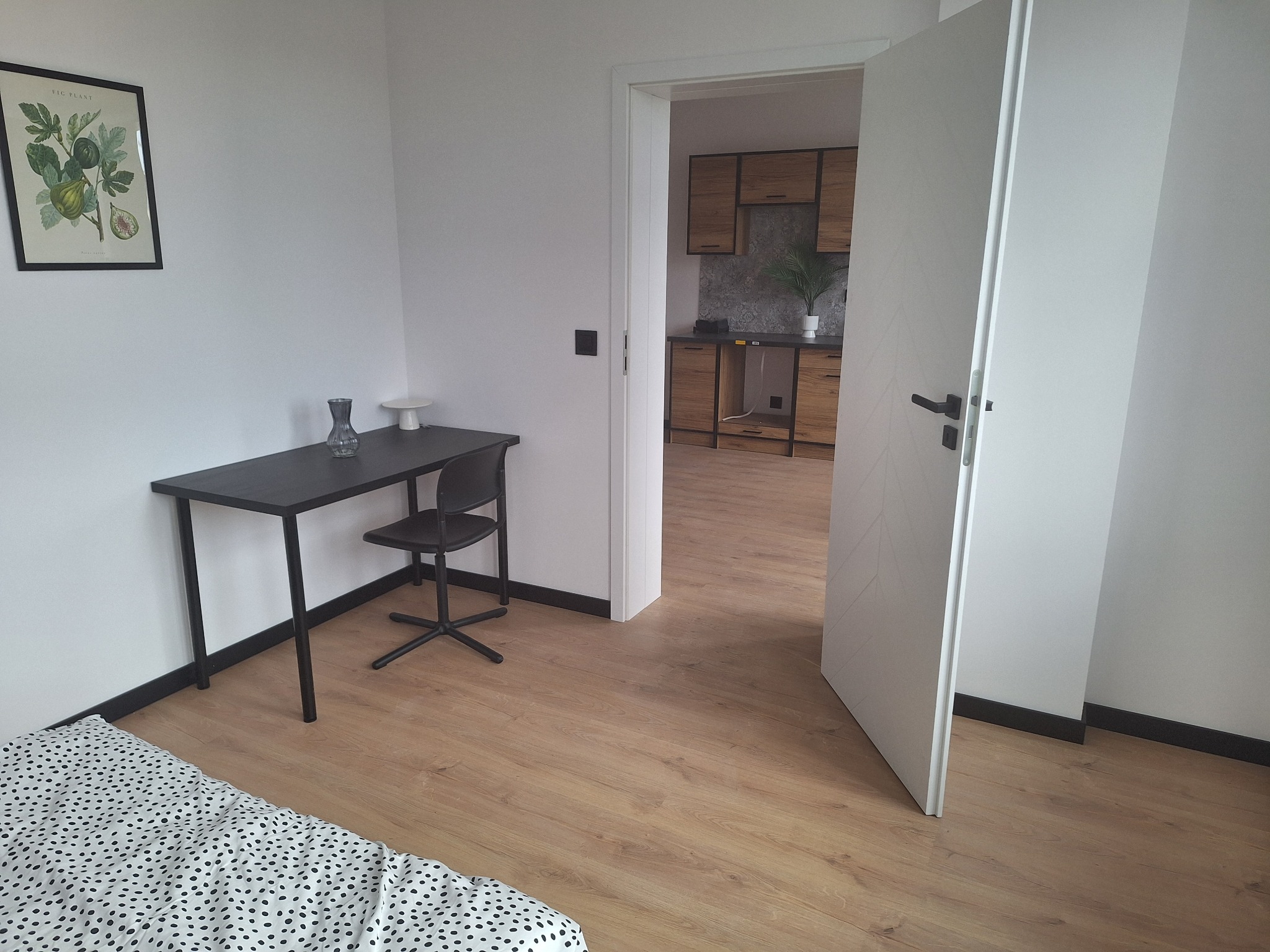 Jasne wnętrze: drewniana podłoga, białe ściany, otwarte drzwi z widokiem na kuchnię. Minimalistyczne biurko z krzesłem i dekoracje. Nowoczesny design.