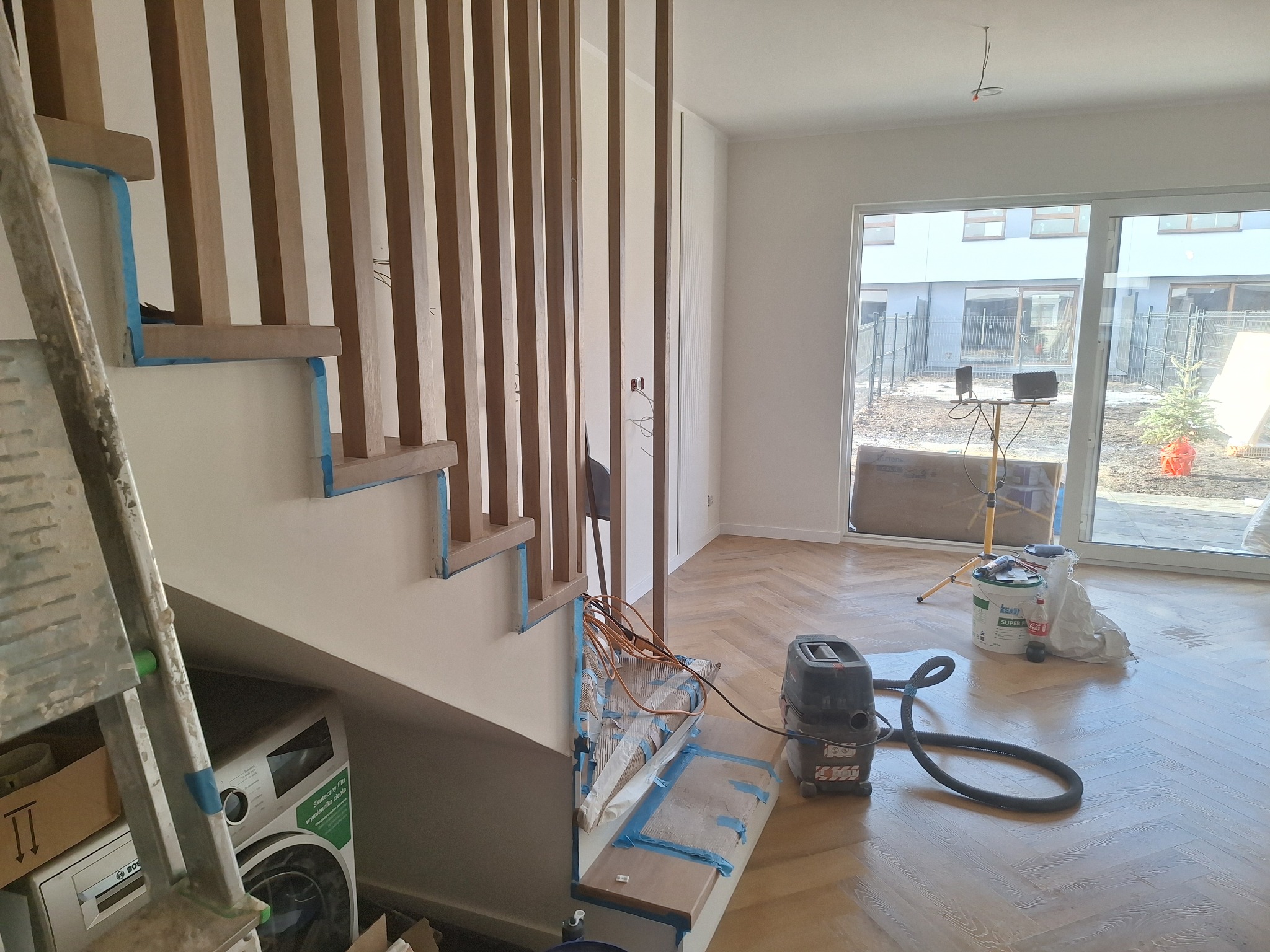 Drewniane schody z barierką w trakcie malowania, zabezpieczone taśmą malarską. Na podłodze parkiet w jodełkę, widoczny odkurzacz przemysłowy i drabina.