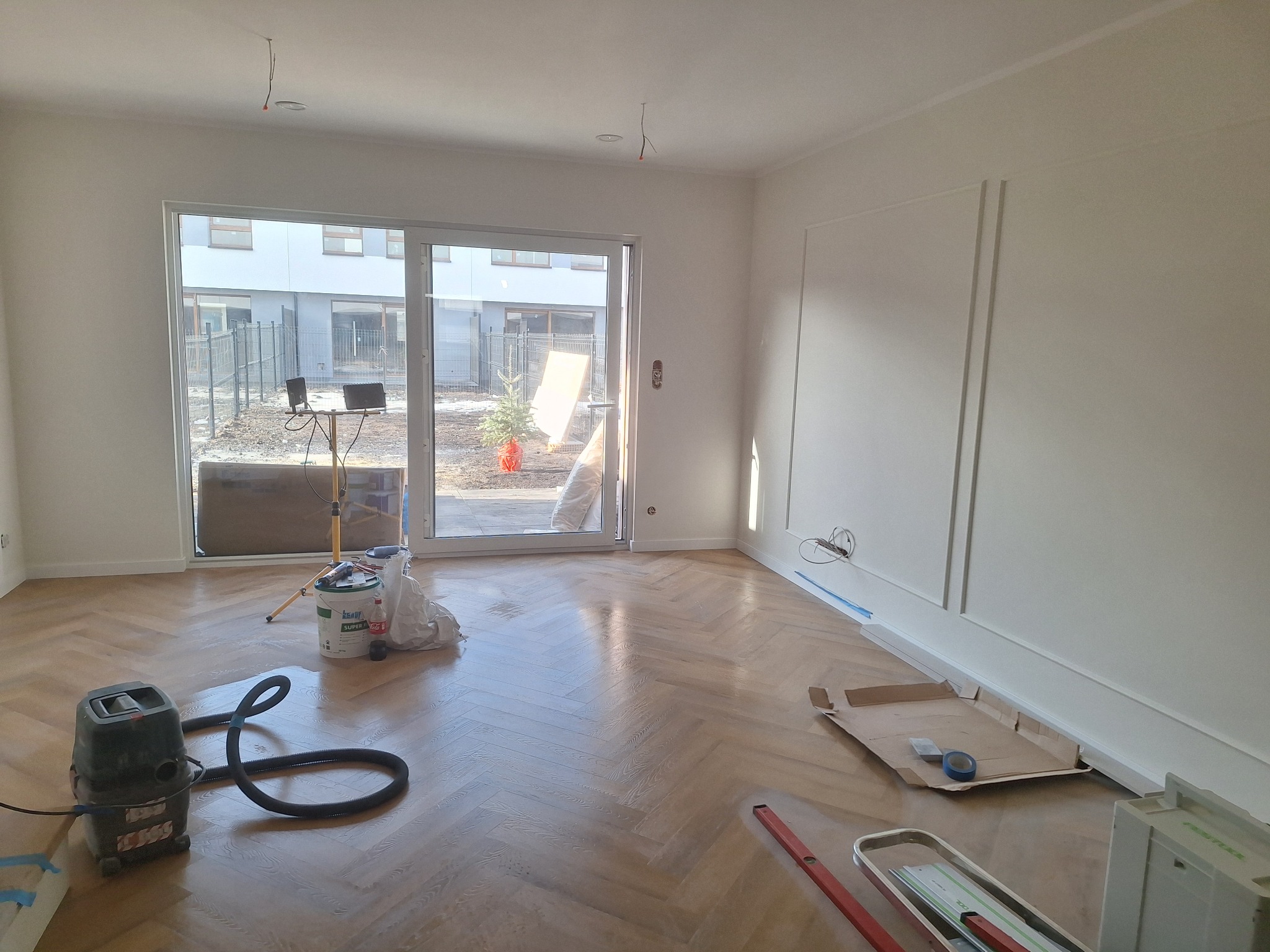 Jasny parkiet ułożony w jodełkę, po lakierowaniu, z widocznym sprzętem do czyszczenia i narzędziami, w przestronnym wnętrzu z białymi ścianami i dużym oknem.