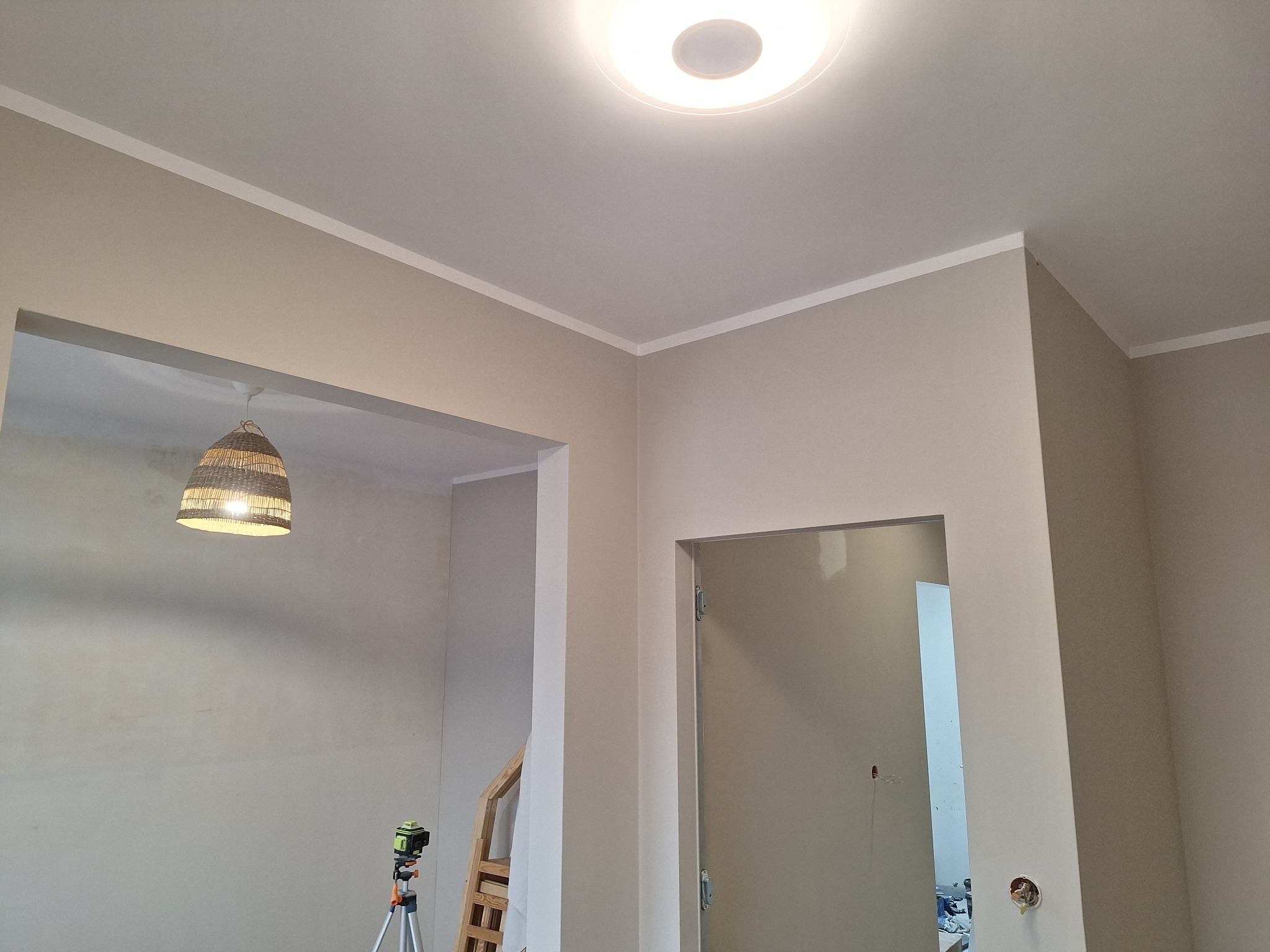 Wnętrze po malowaniu ścian na beżowo z białym wykończeniem sufitu. Widoczny laser krzyżowy, lampa wisząca i fragment drabiny.