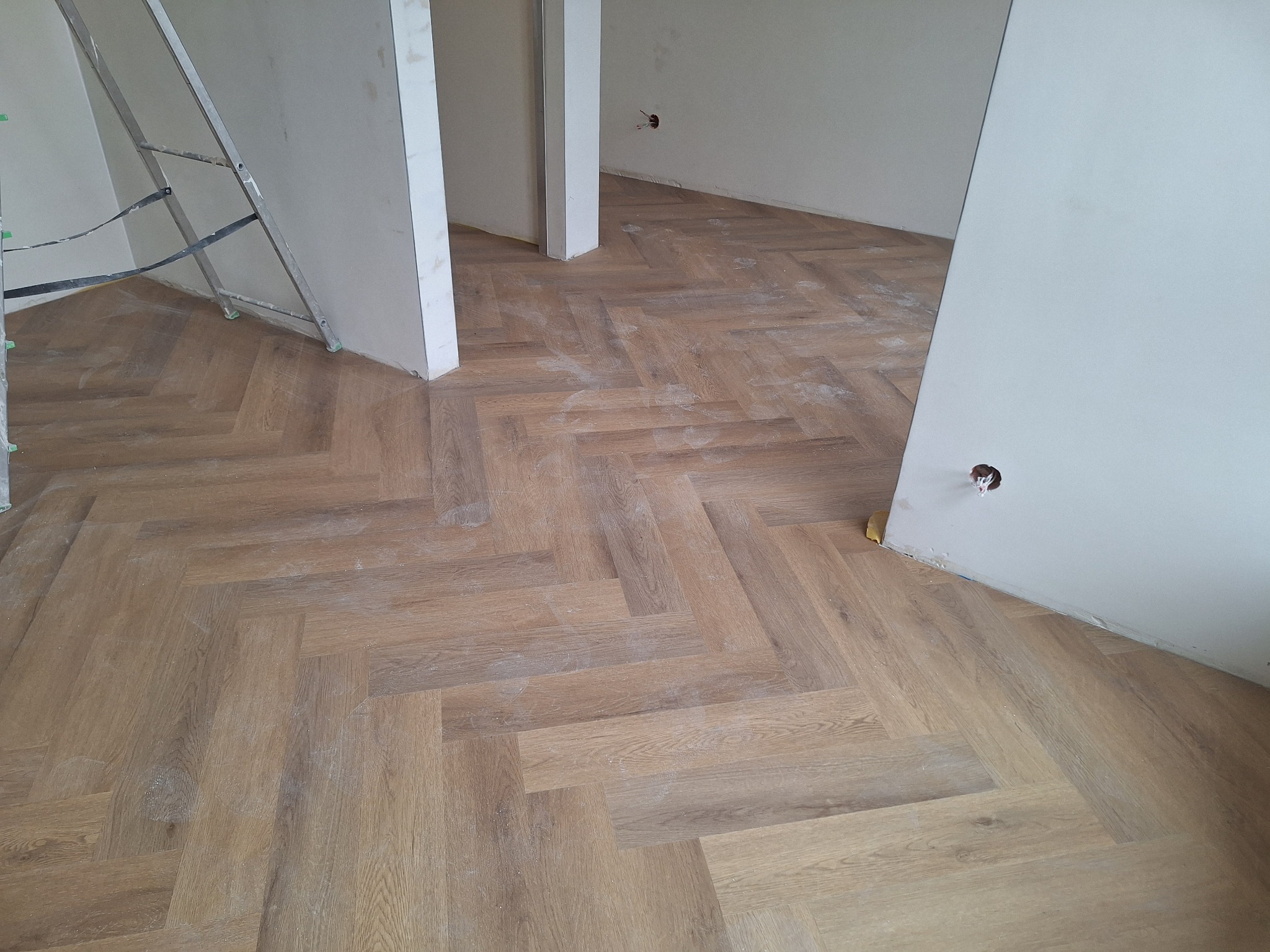 Ułożony parkiet w jodełkę francuską w jasnym odcieniu drewna, widoczna drabina malarska i fragment ściany w trakcie prac wykończeniowych.