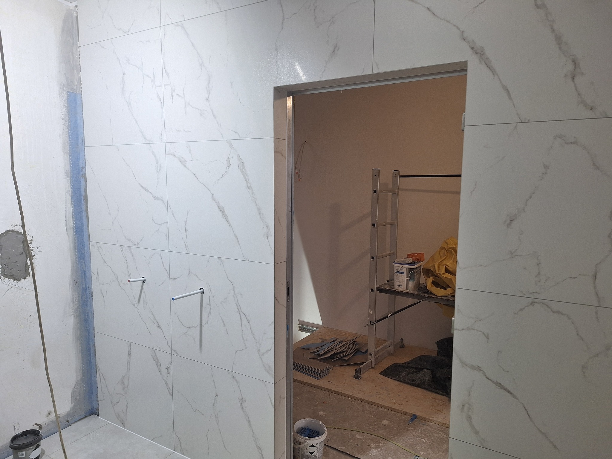 Białe płytki imitujące marmur na ścianie łazienki w trakcie wykańczania. Widoczne wystające rurki i fragment ściany bez płytek. W tle drabina i narzędzia.